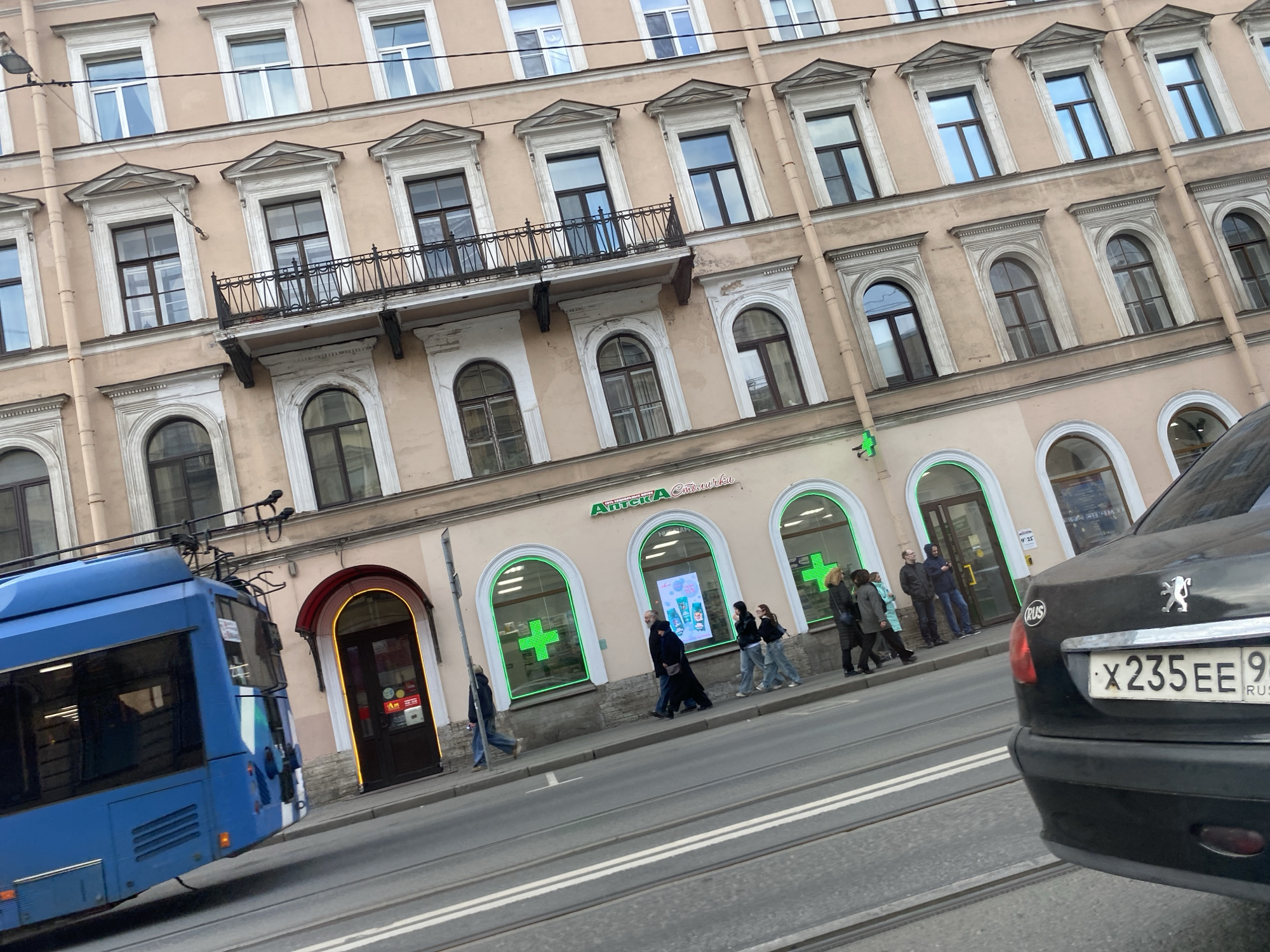 The image depicts a street scene in Saint Petersburg, Russia. A multi-story building with light brown facade is the backdrop, featuring classical architectural details like arched windows and a balcony. A pharmacy called "Apteka" in Russian is located on the ground floor, indicated by green crosses in the window. Pedestrians are walking on the sidewalk in front of the pharmacy. A blue bus is visible on the left side of the frame. A black car, partially in the foreground, has a Russian license plate reading "X235EE 98". Tram tracks are seen on the street.