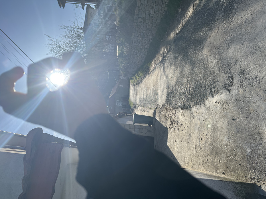 An outdoor scene in Sedlartsi, Bulgaria, depicts a clear, sunny day. In the immediate foreground, a person's left hand is silhouetted, positioned to frame the intensely bright sun, which emits pronounced lens flares through an aperture formed by the fingers. To the left, a portion of a building's reddish-tiled roof and gutter is visible. A narrow, partially paved road or path recedes into the midground. Adjacent to the road on the left stands a dark green wheeled trash bin next to a stone-clad building wall. On the right side of the road, a dry-stacked stone retaining wall supports an elevated grassy embankment. Further down the path on the left, a house with a reddish roof and a visible white-framed window is situated. On the right, a multi-story building with a light-colored roof appears on higher ground, partially obscured by bare tree branches. Multiple power lines traverse the clear blue sky overhead.