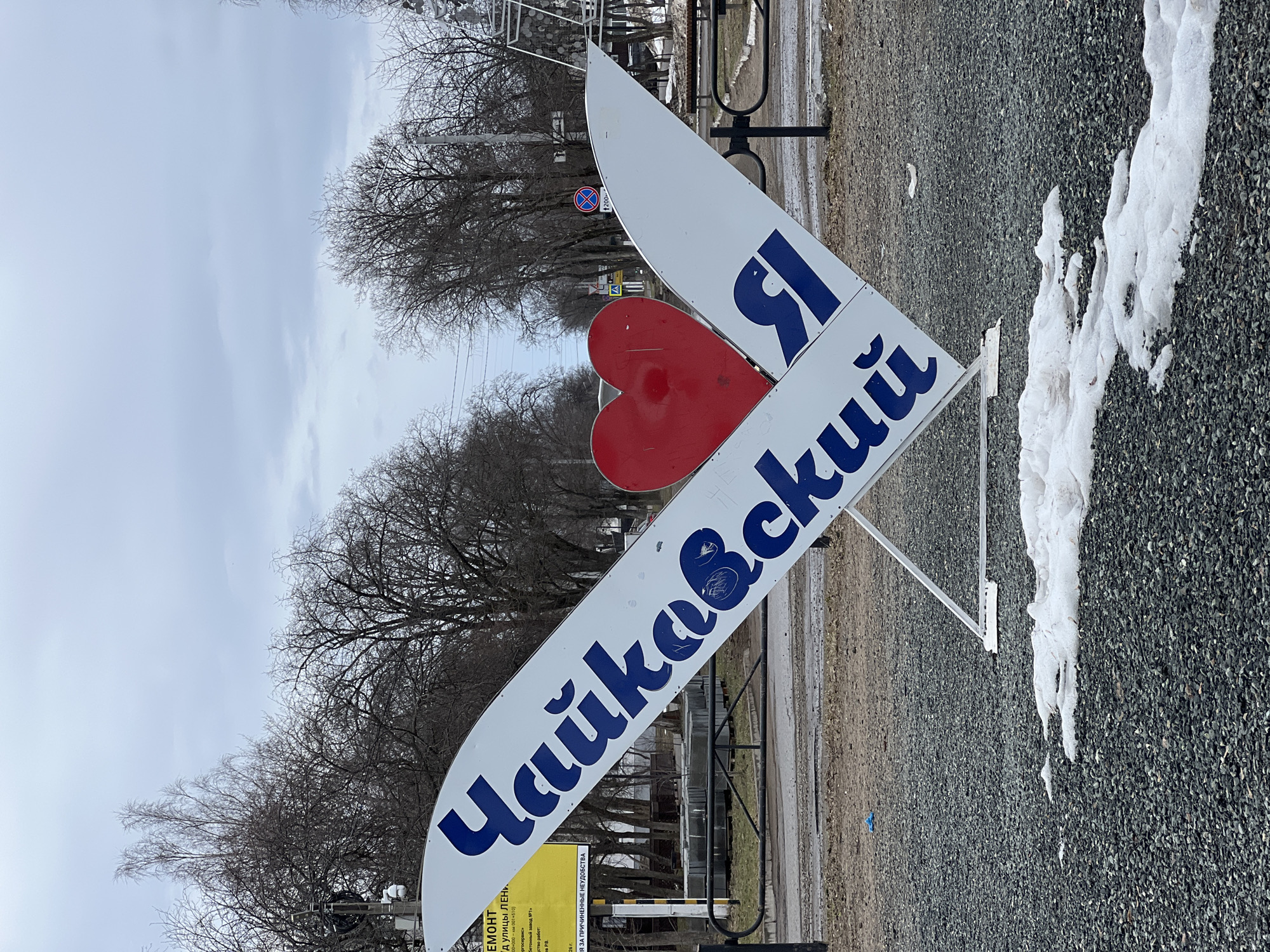 The image captures a roadside sign in Chaikovsky, Russia. The sign is designed to resemble an "I love" graphic, where the word "love" is replaced with a red heart, and the city name "Chaikovsky" is written in Cyrillic script. A single letter "Я" is placed on the top part of the sign near the heart. The sign is situated beside a patch of gravel and melting snow. In the background, there is a row of bare trees lining a road, with some pedestrian railing visible behind the sign. A distant blue road sign is seen through the trees. A sign with fine print is also nearby, possibly giving information or directions. The overall scene appears to be taken in a public space within the city, likely on a day with overcast skies.
