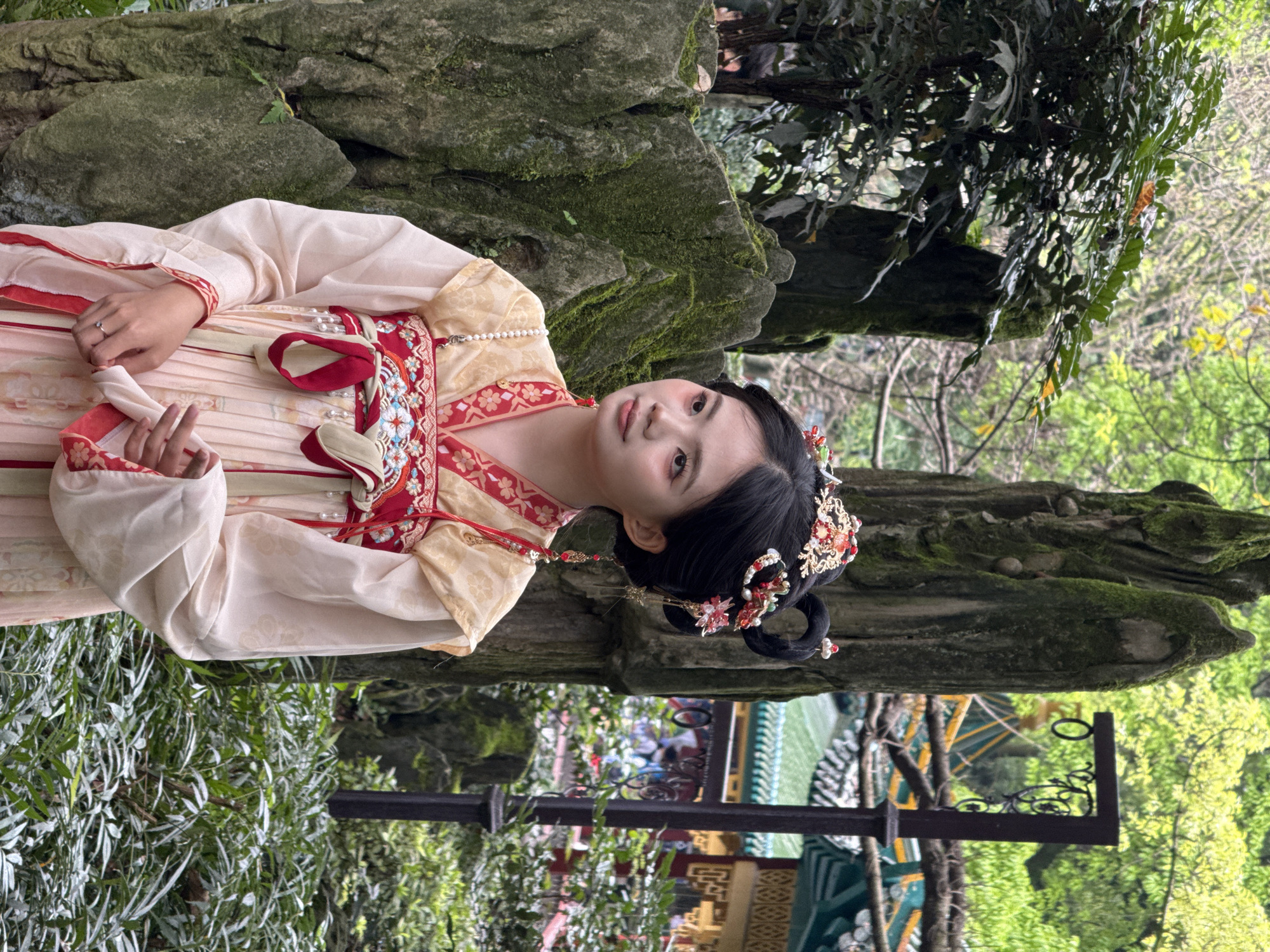A woman in traditional Hanfu attire is posed outdoors in a garden-like setting. The woman has black hair styled in a traditional updo, adorned with floral ornaments. She is wearing a cream and peach-colored robe with intricate red and gold embroidery. She looks towards the camera. She is standing next to a rock covered in moss. The background consists of lush greenery, trees, and a glimpse of a traditional Chinese architectural structure with a green tiled roof and decorated eaves. This photo was taken in Yuzhong District, China.