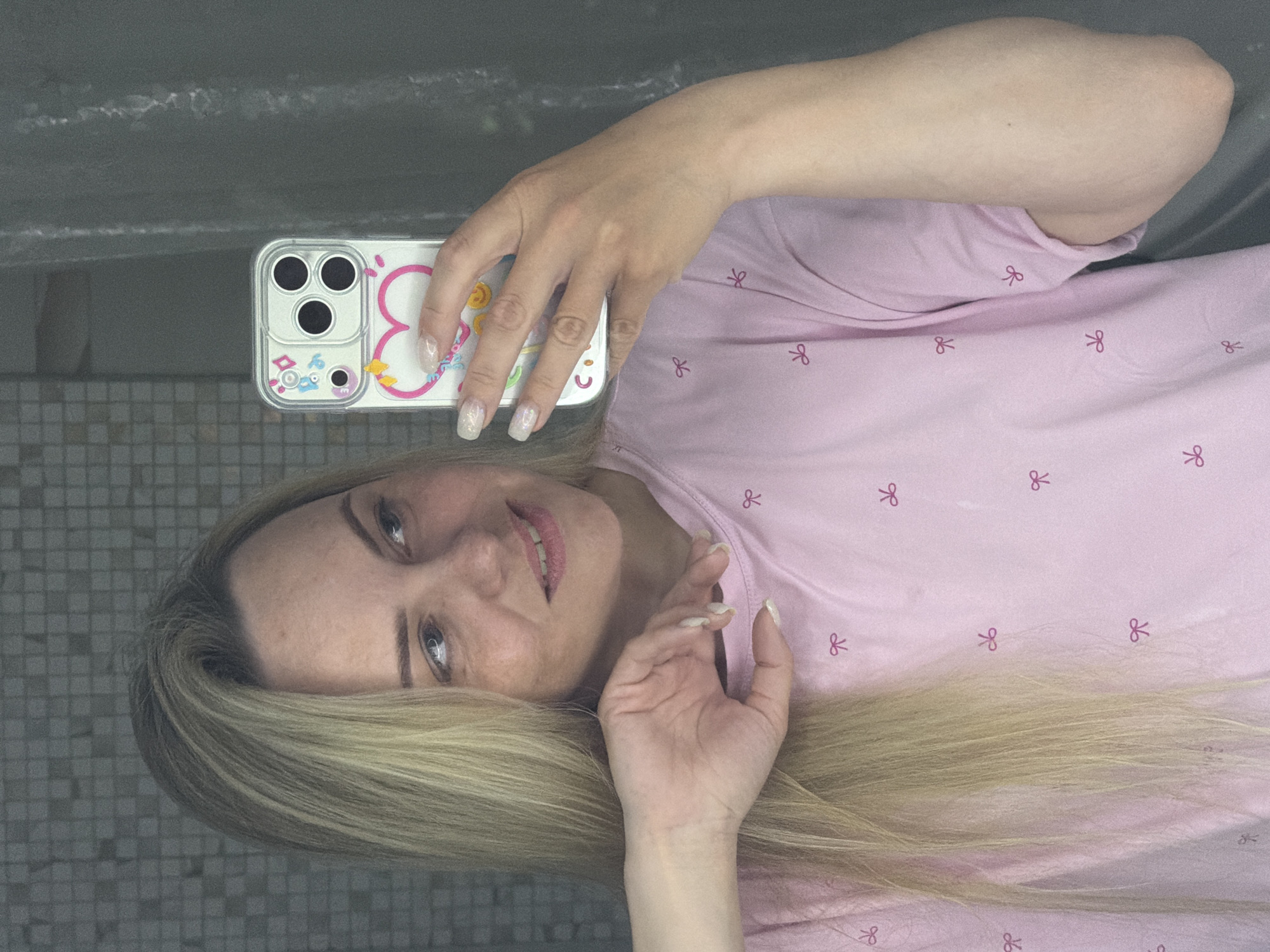 A woman is holding a smartphone with a decorative case, taking a selfie. The location is not provided in the image and cannot be deduced. The woman has blonde hair, blue eyes, and is wearing a pink shirt with a bow pattern. She is indoors. The background appears to be a wall or tiled surface.