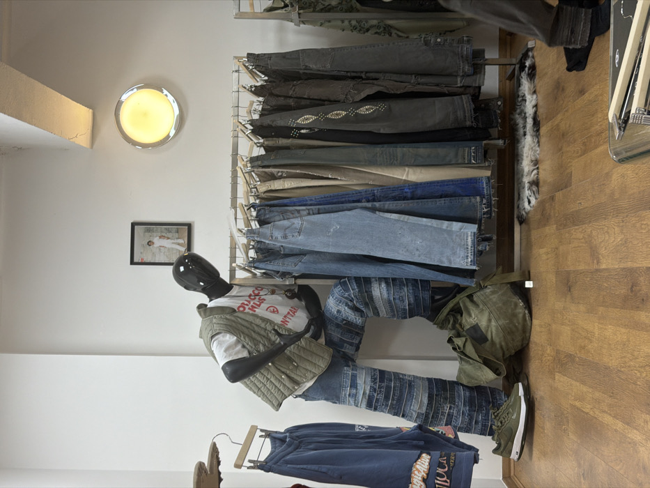 Interior view of a retail space in the city of London, United Kingdom. The scene features a glossy black mannequin in a dynamic, crouching pose, positioned on the left side. The mannequin is dressed in a white t-shirt with red text (partially visible, reading "TOBACCO KILLS... DON'T START"), an olive green quilted vest, and wide-leg denim jeans constructed from various patches of light and dark blue denim. A single olive green Nike Air Force 1 sneaker is visible on its left foot.
On the light brown wooden floor, immediately below the mannequin, lies a large, olive green canvas duffel bag. To the right, a metal clothing rack holds approximately 20 pairs of trousers or jeans in various shades of blue, grey, and black, some featuring distressing or embellishments like silver studs. Beneath this rack, a small, multi-toned fur rug rests on the floor.
The walls are painted white. A rectangular black-framed photograph depicting a person in white clothing hangs on the back wall above the clothing rack. To the right of the photograph, a circular wall-mounted light fixture emits a warm yellow glow. A concrete or painted white beam is visible across the upper right portion of the ceiling. On the far left, another partial clothing rack displays several blue garments.