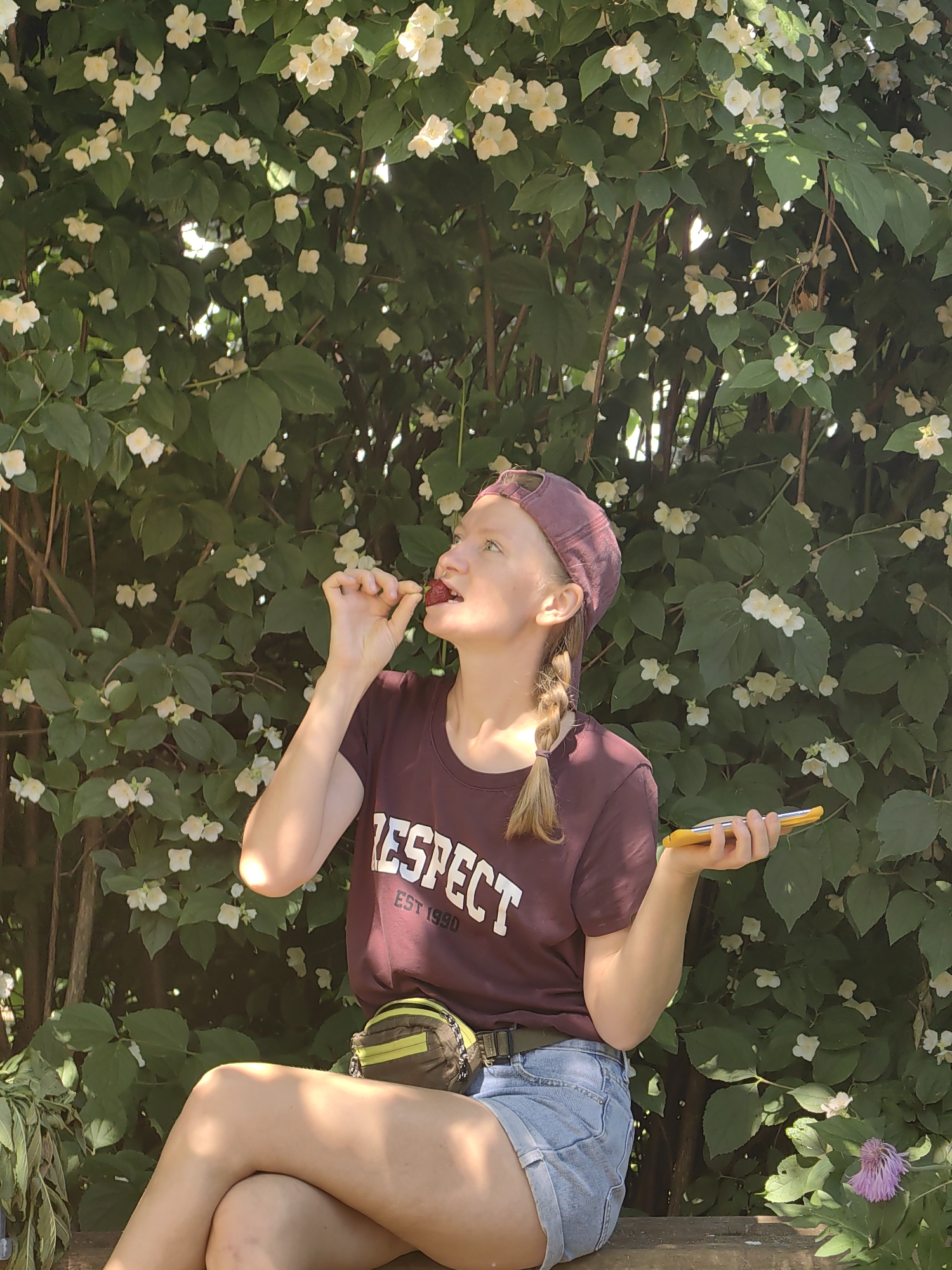 A young woman with blonde hair in a braid wearing a burgundy t-shirt with "RESPECT" printed on it and a burgundy baseball cap sits on a wooden bench in front of a bush with white flowers. She is eating a strawberry and holding a yellow phone in her right hand. She is wearing denim shorts and a fanny pack around her waist. The background is filled with greenery and sunlight is dappling through the leaves.
