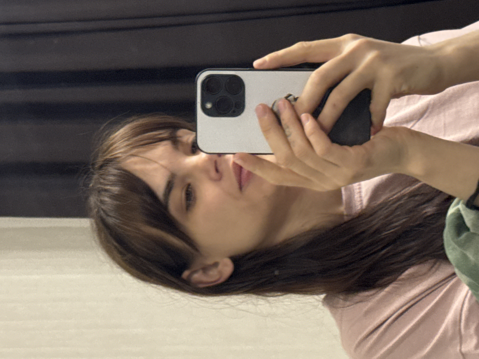 A person with shoulder-length brown hair is holding a white smartphone, obscuring part of their face. The phone has a triple-lens camera system visible on the back. The person is wearing a light pink top, and a small portion of a green garment is visible. The background features a reflective surface, possibly a mirror or a glossy wall, with dark horizontal lines. There is not enough information to discern the location or provide details on specific actions or interactions.