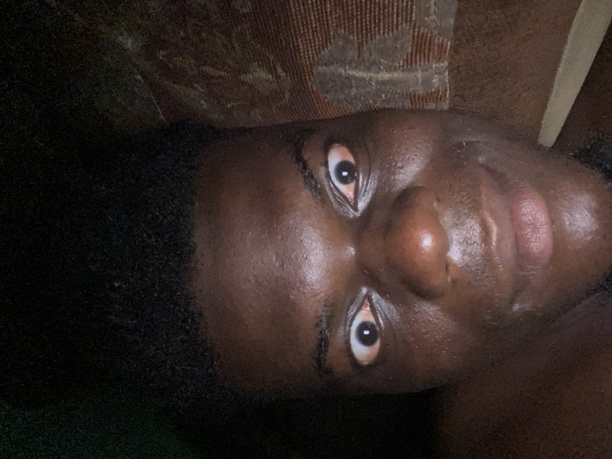 A close-up, slightly angled view of a person's face, likely of African descent. The person is facing upwards with their eyes open. The background includes what appears to be patterned upholstery or a textured surface. Part of the scene is shrouded in darkness, obscuring detail. The location and city are unknown.