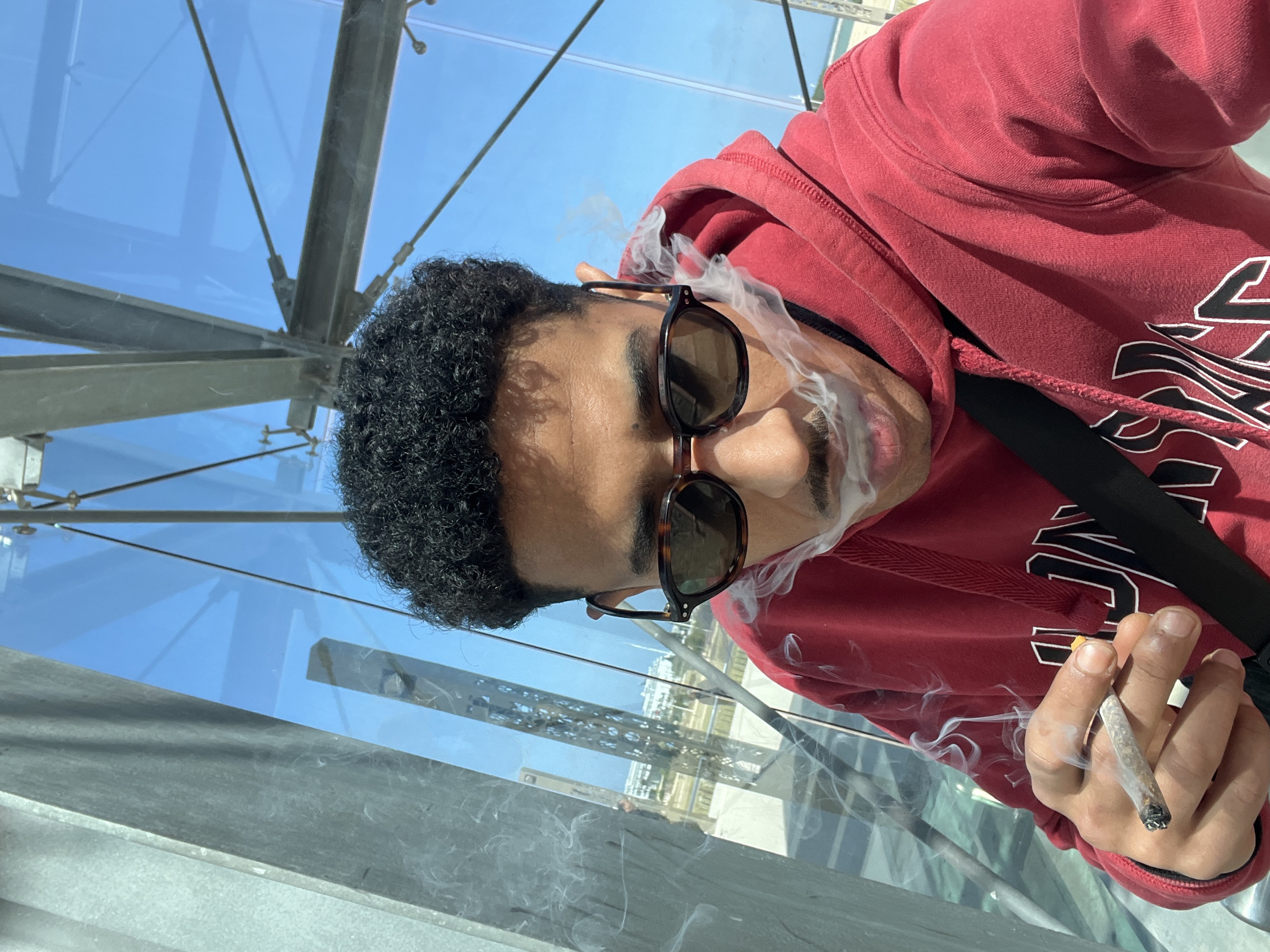 A male individual is depicted in a close-up, selfie-style photograph, positioned within a modern architectural structure composed of glass panels and steel beams under a clear blue sky. The individual, appearing to be a young adult with dark, curly hair, a moustache, and light brown skin, wears dark, possibly tortoise-shell framed sunglasses and a red hooded sweatshirt with partial white and black graphics. A black strap crosses his chest. His right hand holds a lit, hand-rolled cigarette, emitting smoke. Smoke is also seen exhaling from his mouth and nostrils. The background, viewed through the transparent glass, reveals a coastal urban environment in Tanger, Morocco, featuring several buildings, some appearing white or light-colored, a distinct lattice-patterned pillar, and a distant body of water, indicative of a promenade or marina area along the city's waterfront.