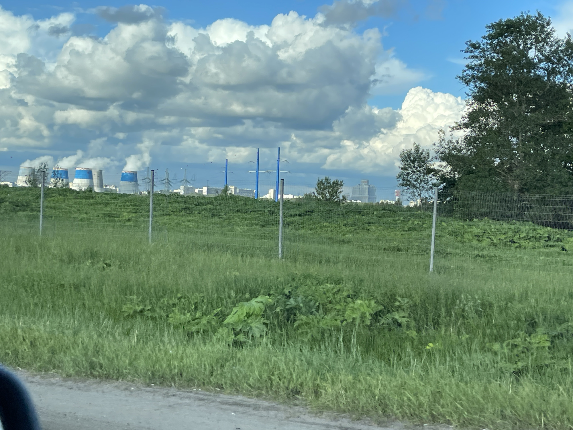 The image captures a view from the outskirts of Novodrozhzhino, Russia. A field of green grass occupies the foreground, with a wire fence running across it. In the background, several industrial cooling towers with blue and white markings are visible. High-voltage power lines and additional buildings suggest an industrial area or power plant in proximity to the town. A stand of trees is located on the right side of the image. The sky is partly cloudy with a gradient from light blue to white.