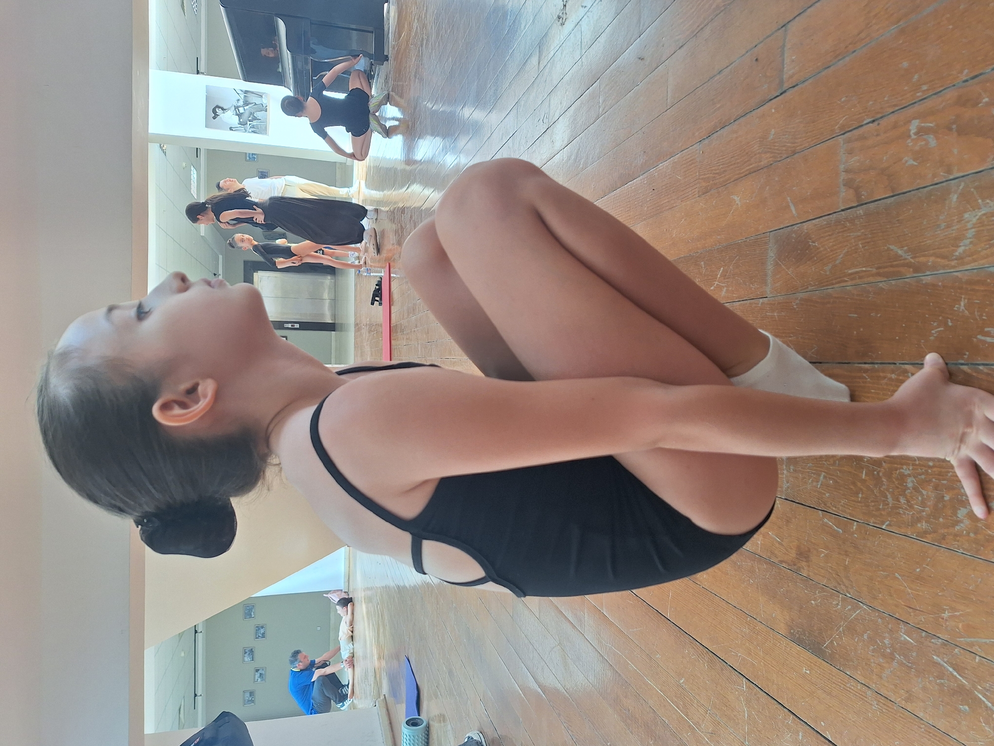 In a dance studio with wooden floors, a young girl in a black leotard is practicing a yoga pose. Her legs are drawn up to her chest, and her arms are wrapped around them. Her head is tilted back, and her eyes are closed, suggesting concentration or relaxation. In the background, reflected in a large mirror, several other dancers are also practicing. Some are standing, while others are in similar poses. A piano is visible to the left, and a few framed pictures hang on the wall. A man in a blue shirt and dark pants is seen helping a young boy with a pose. The lighting suggests it is daytime.