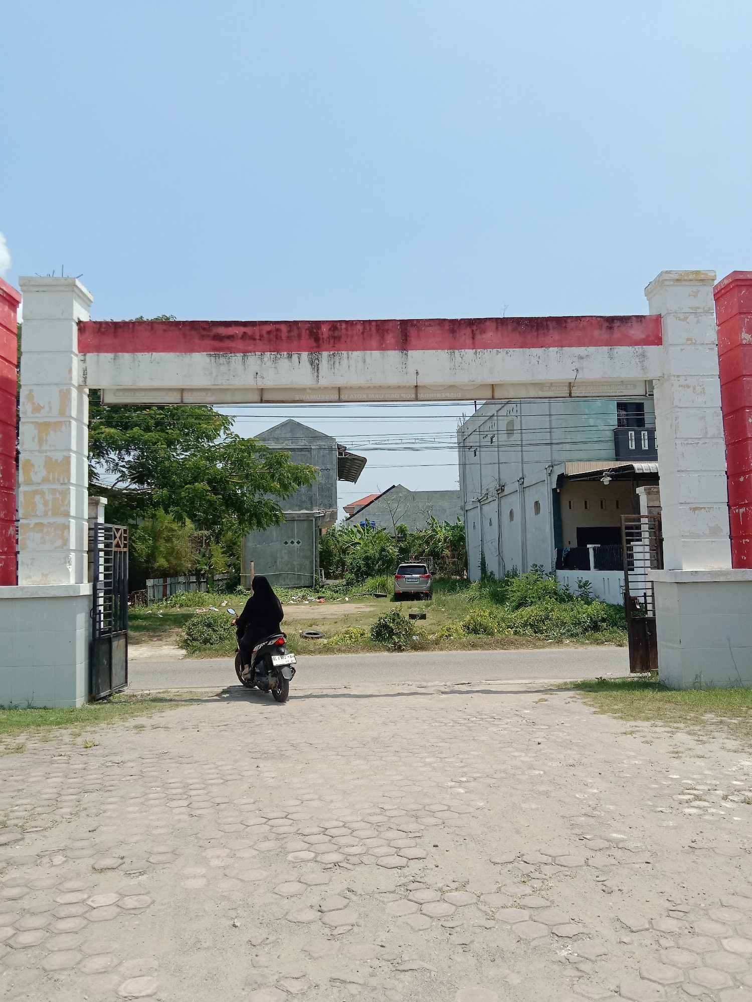 The image depicts a bright, sunny outdoor scene in Lhokseumawe, Indonesia, centered around a large, weathered gateway. A person, dressed in black and wearing a head covering, is seen from behind riding a dark scooter with the license plate "DC 6292 XBH". They are positioned on a paved road, either entering or exiting the area through the gateway.
The gateway itself is a prominent structure composed of two large pillars and a horizontal beam, all painted in a faded and peeling red and white, reminiscent of the Indonesian flag. The paint is significantly chipped and discolored, showing signs of age and exposure. Faint, worn text is visible on the underside of the horizontal beam, appearing to be an advertisement for rent in Indonesian, with discernible words such as "DISEWAKAN" (for rent) and "SEWAKAN KOTAK SEWA". Metal gates are attached to the inner sides of the pillars.
Beyond the gateway, the background features several modest, two-story buildings, some appearing residential or unfinished, interspersed with abundant green trees and overgrown vegetation. A silver car is parked on the left side of the road in the distance. The foreground consists of a forecourt paved with hexagonal concrete blocks that are uneven, dusty, and partially covered with sand and weeds. The sky above is clear and bright blue, suggesting midday or early afternoon. The scene conveys an impression of a somewhat unmaintained or developing area under clear weather conditions.