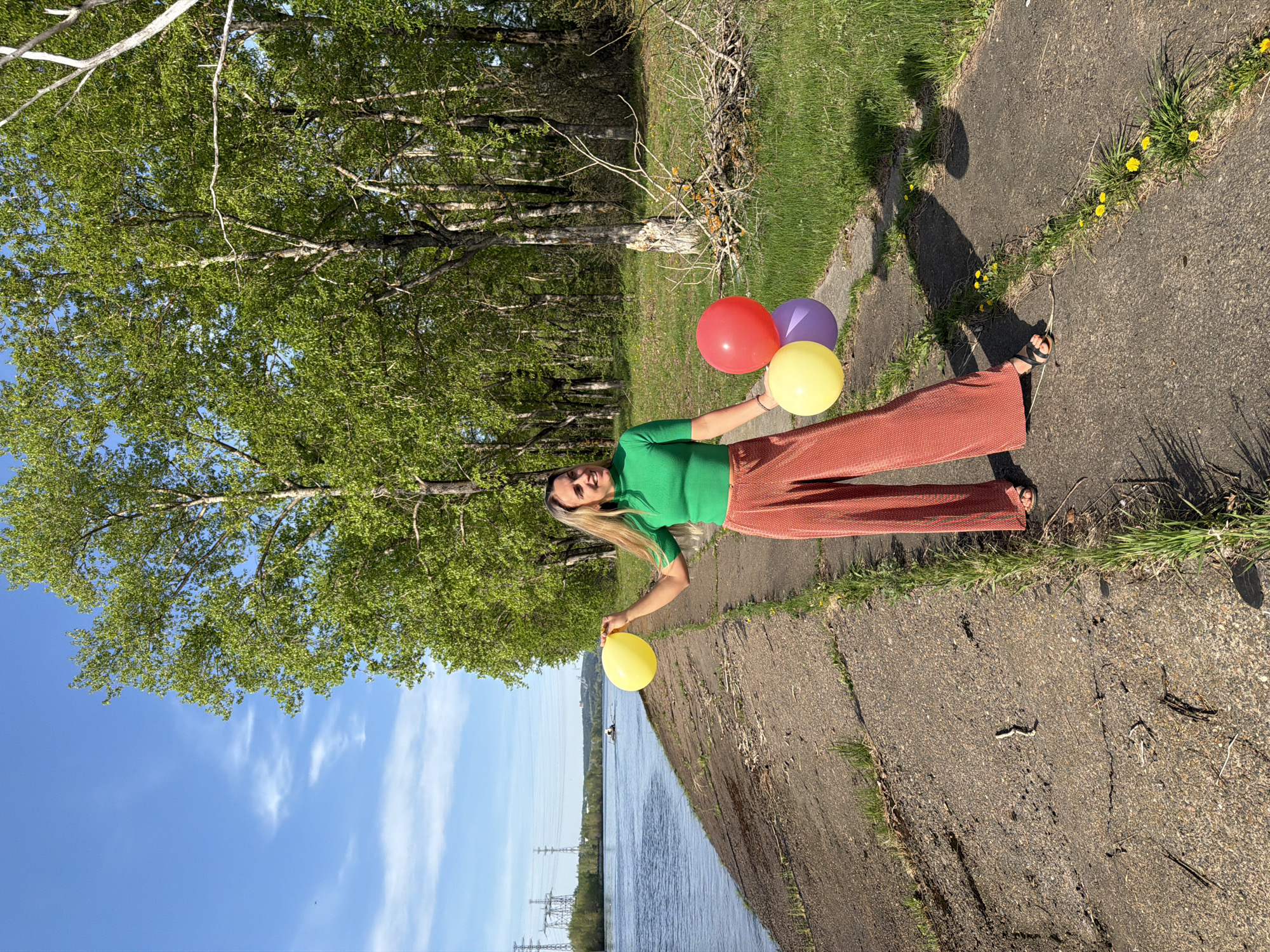 A woman stands on a concrete path next to a body of water in Chaikovsky, Russia. She is holding balloons and wearing a green shirt and rust-colored pants. Trees are behind her, and power lines are visible across the water.