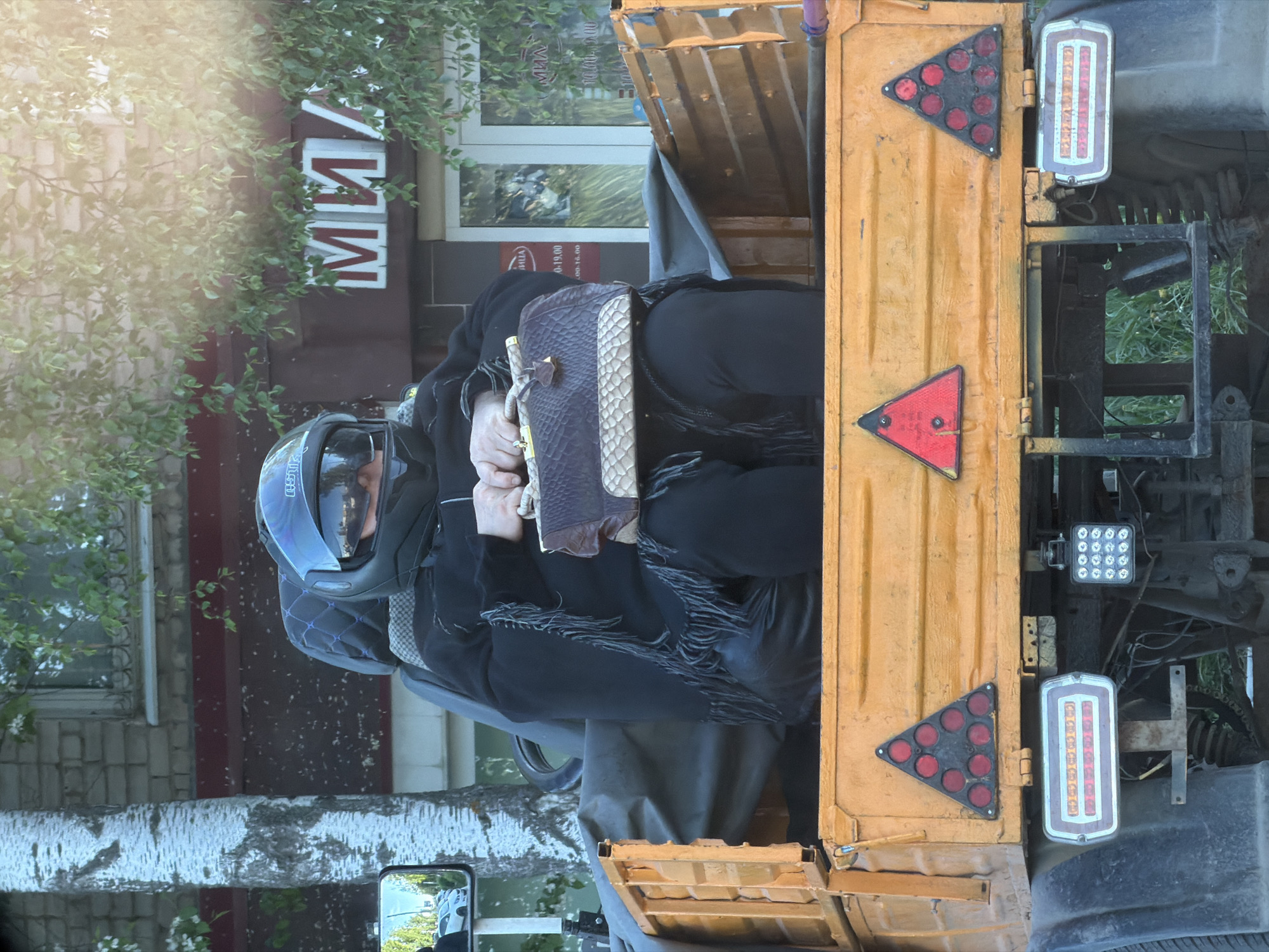 A person in a motorcycle helmet and dark clothing is sitting in the back of a yellow truck. They are holding a handbag in their hands. The truck has triangular reflectors and lights on the back. The setting appears to be in the city of Chaikovsky, Russia, based on the Cyrillic text on a building in the background. The background also includes trees and a building.