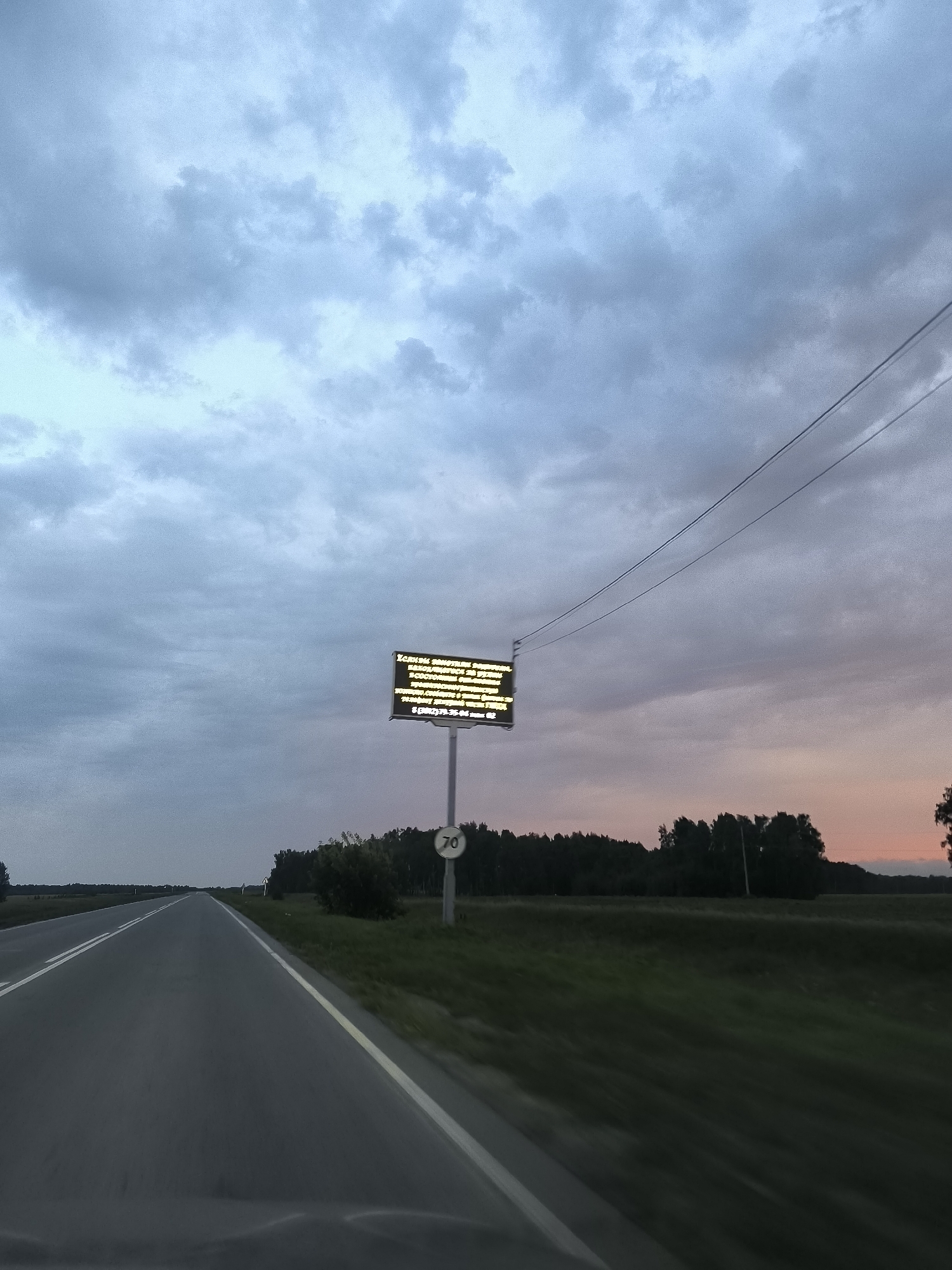 The image shows a road leading into the distance under a cloudy sky. On the right side of the road, there is a digital sign with text in Russian, and a speed limit sign indicating 70. The surrounding landscape consists of fields and trees. The overall scene appears to be on the outskirts of a rural area, potentially near Avly, Russia, given the context. The lighting suggests it is either dawn or dusk.