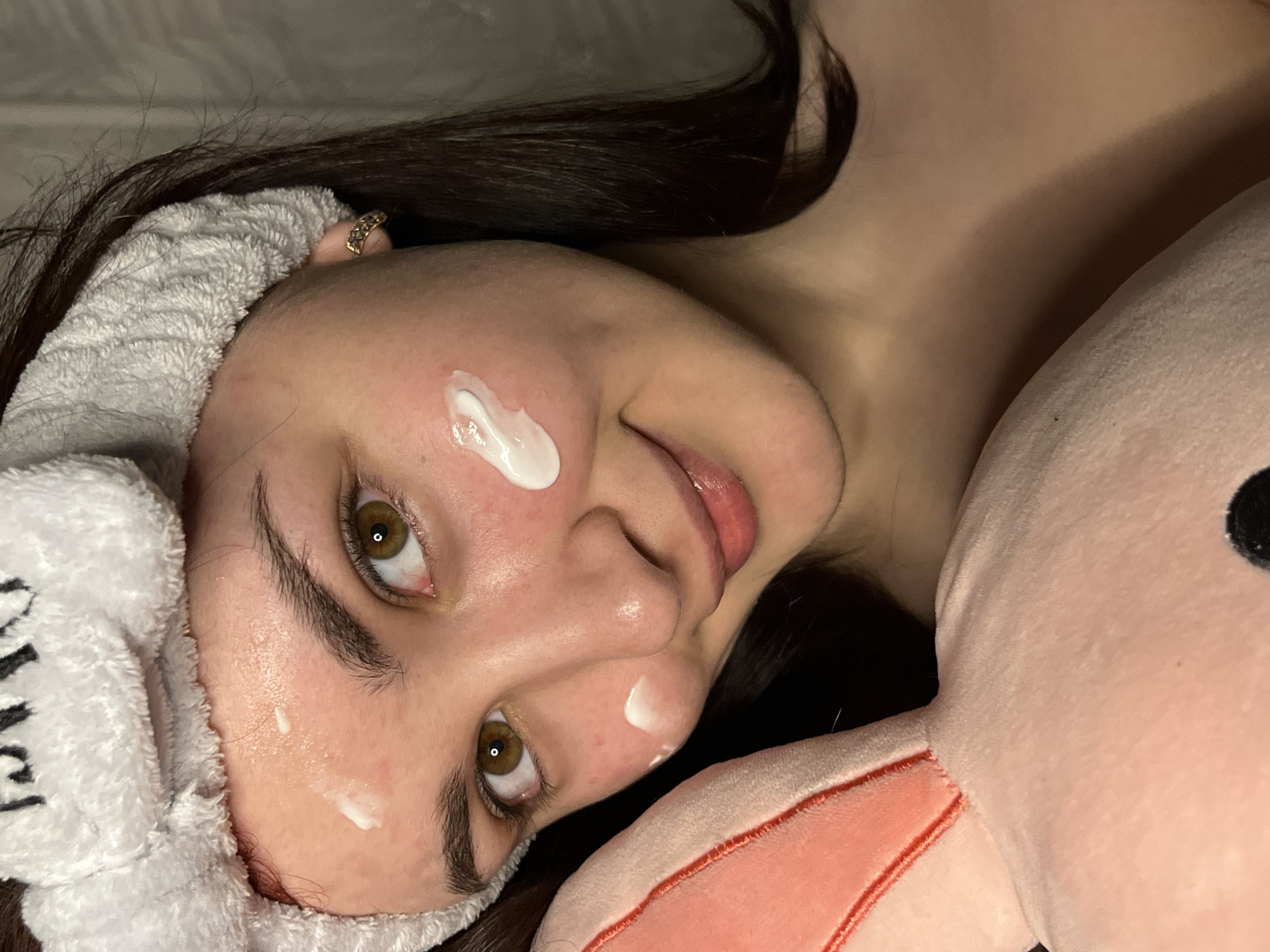 A woman with dark hair and hazel eyes is lying down with a pink plush rabbit beside her. She is wearing a white terry cloth headband and has three small dabs of white cream on her face: one on her left cheek, one on her nose, and one on her right cheek near the headband. She is looking directly at the camera. A gold ring is visible on her finger. The background appears to be a light-colored surface. There is no information visible in the image to place the location as the city of Neftekamsk, Russia.