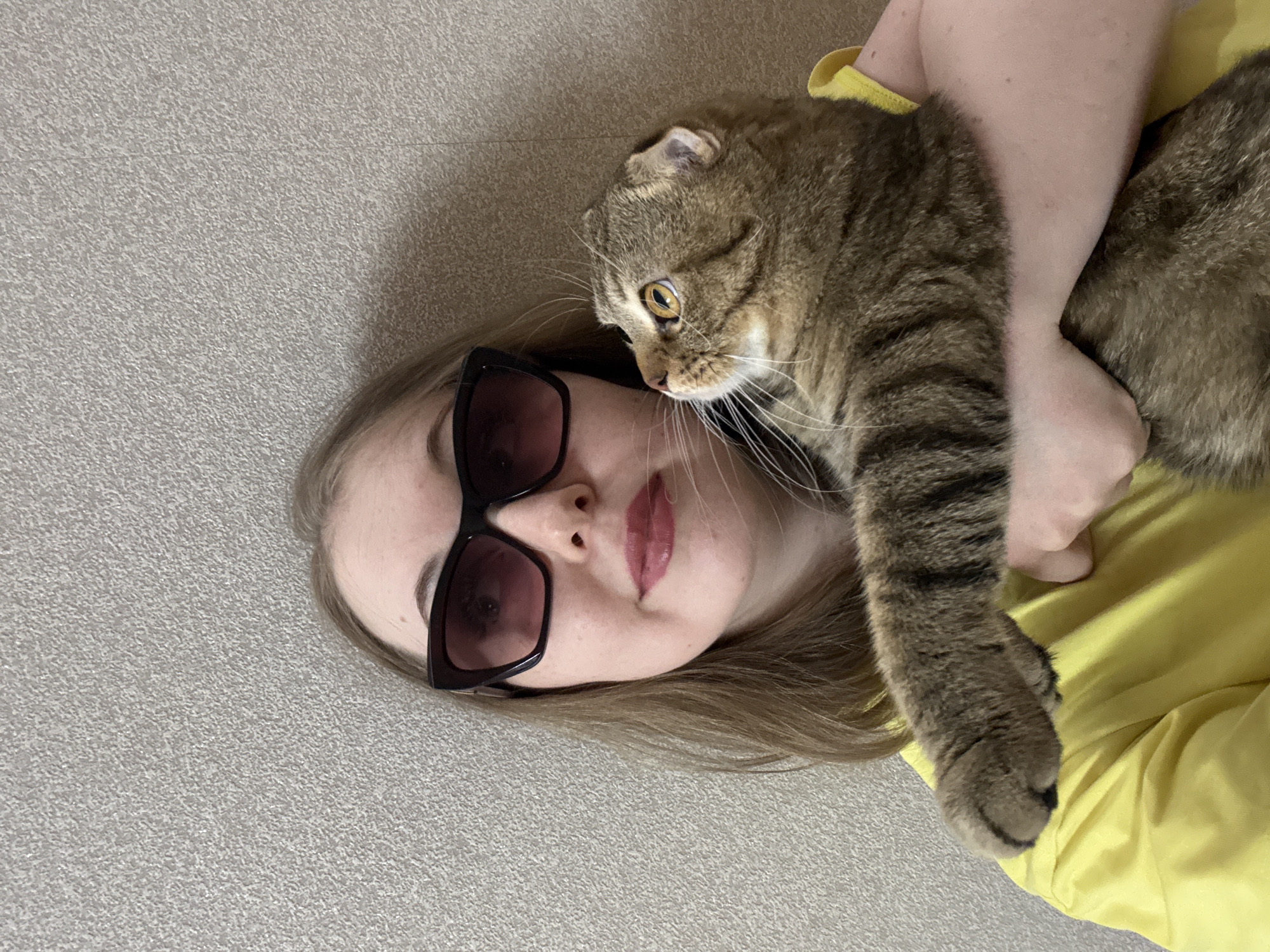 This image captures a close-up, overhead view of a young woman interacting with a tabby cat in what appears to be an indoor setting. The woman is lying down, with her head resting on a textured light grey surface, possibly a carpet or soft flooring. She has light brown hair, is wearing stylish dark rectangular sunglasses, and a bright yellow top or shirt. A hint of light pink lipstick is visible on her lips.
A brown and black striped tabby cat is comfortably resting on her upper chest or shoulder, with its body draped across her arm. The cat, possibly a Scottish Fold given its distinctive folded ears, has prominent white whiskers and large, round, amber-colored eyes that are fixed intently towards the left side of the frame. One of the woman's arms is visible, gently supporting the cat, indicating a tender interaction. The scene conveys a sense of affectionate companionship and quiet relaxation between the woman and her feline companion. The lighting is soft and ambient, characteristic of an indoor environment. No text is visible in the image.