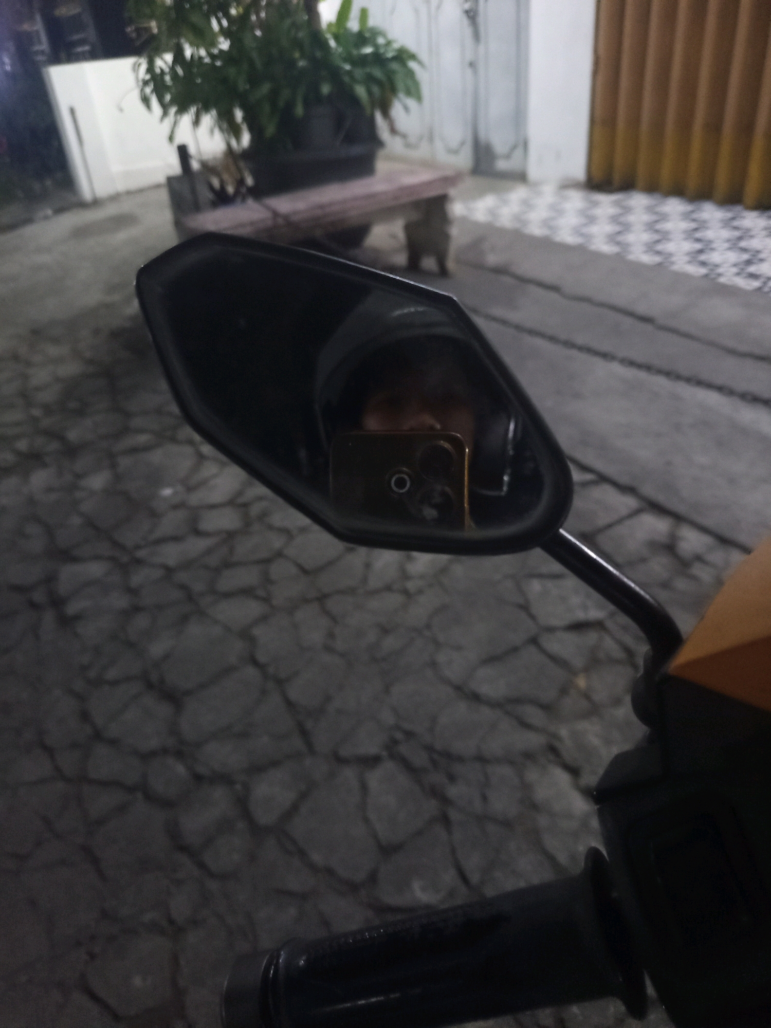 A motorcycle's side-view mirror reflects a person holding a gold smartphone. The person is wearing a dark helmet or head covering, and only their eyes and forehead are visible. The background in the mirror shows an outdoor setting at night, possibly a paved area with a patterned tile walkway and a building with brown shutters. The mirror itself is mounted on the motorcycle's handle. The overall scene suggests a casual setting, perhaps in front of a residence or a business in Singosari, Indonesia, given the commonality of motorcycles in the region.