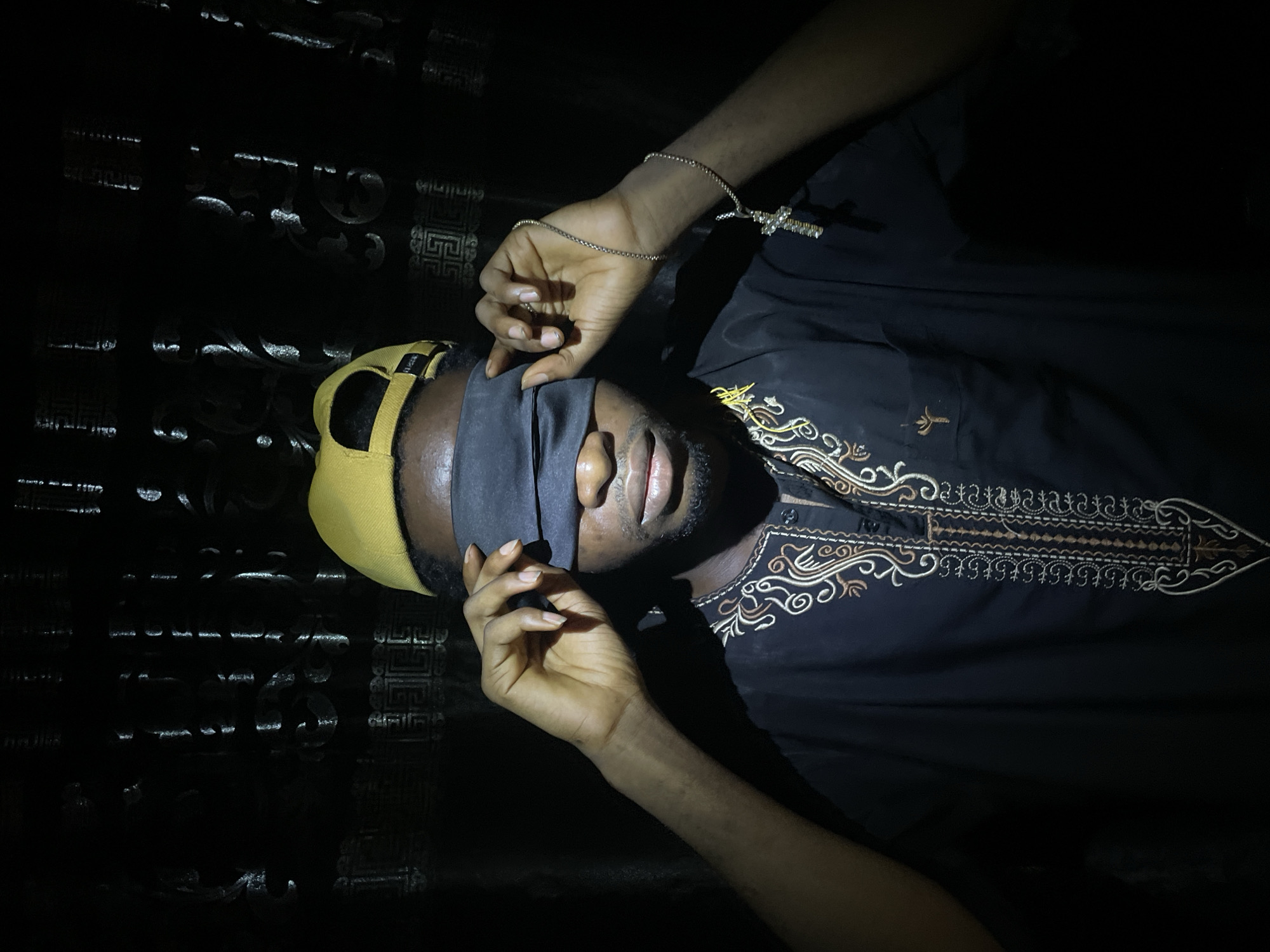 The image features a man lying down, with a black blindfold covering his eyes. He is wearing a yellow baseball cap and a black shirt with ornate embroidery around the collar. His hands are positioned near the blindfold, as if adjusting it. A silver necklace with a cross pendant is visible on his right wrist. The background appears to be a dark surface with a subtle patterned texture. The lighting is dim, casting shadows and creating a sense of intimacy or mystery. There is no explicit information that this photograph was taken in Lokoja, Nigeria.