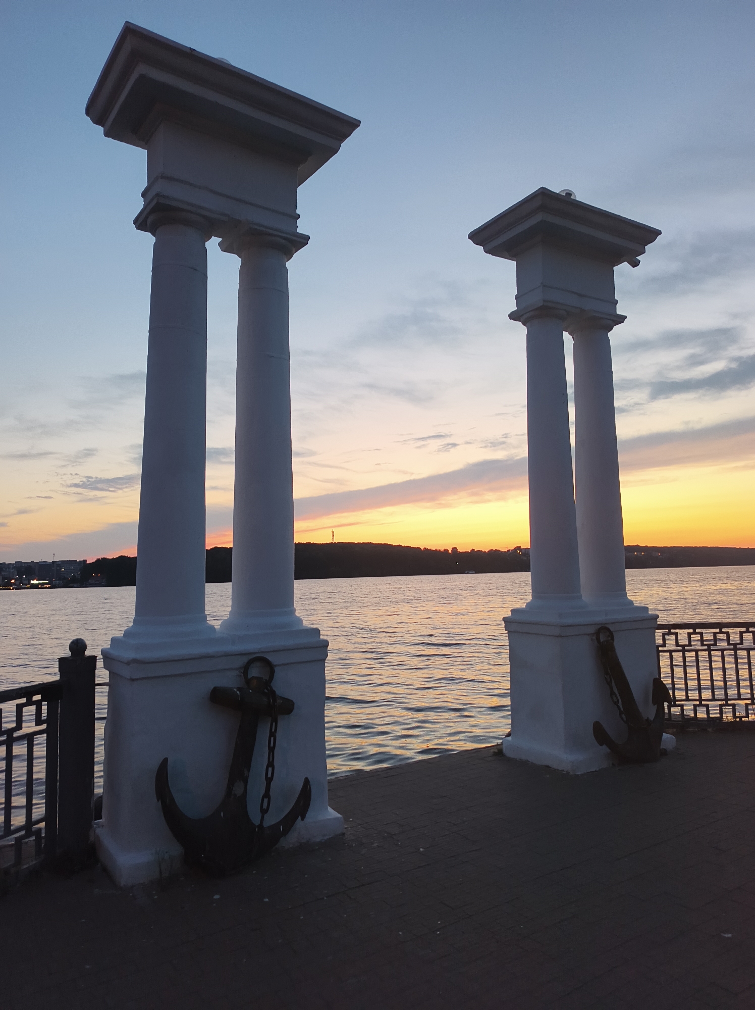 The image shows two classical-style white columns on a paved walkway next to a body of water. Anchors are attached to the bases of the columns. A metal railing runs along the edge of the walkway. The sky is clear with a few clouds, and the sun appears to be setting over a distant treeline and buildings across the water, casting a warm glow. This scene is likely located on the Ternopil Pond embankment in Ternopil, Ukraine, known for its scenic views and recreational areas. There are no people visible in the image.
