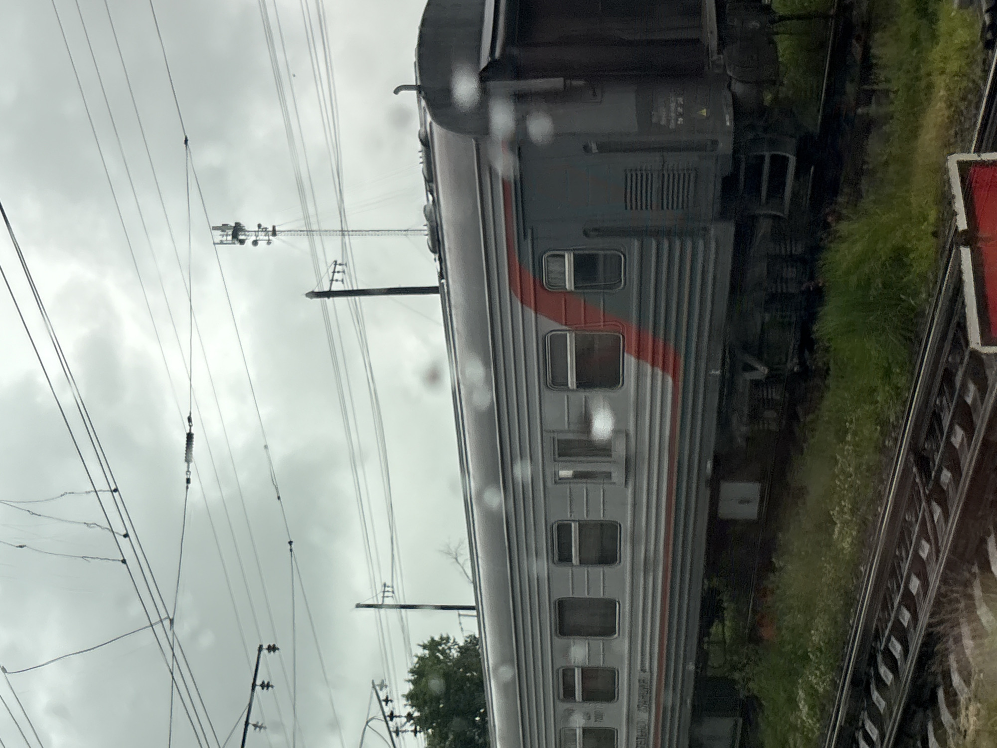 The image depicts a train car of a gray passenger train with a red stripe running horizontally across its side. The train is on a track with overhead power lines. In the background, there is a communication tower, a tree, and a cloudy sky. The foreground shows the train tracks and some green grass. The image appears to be taken from a window, as there are water droplets on the glass. The location is identified as Kotelnich, Russia.