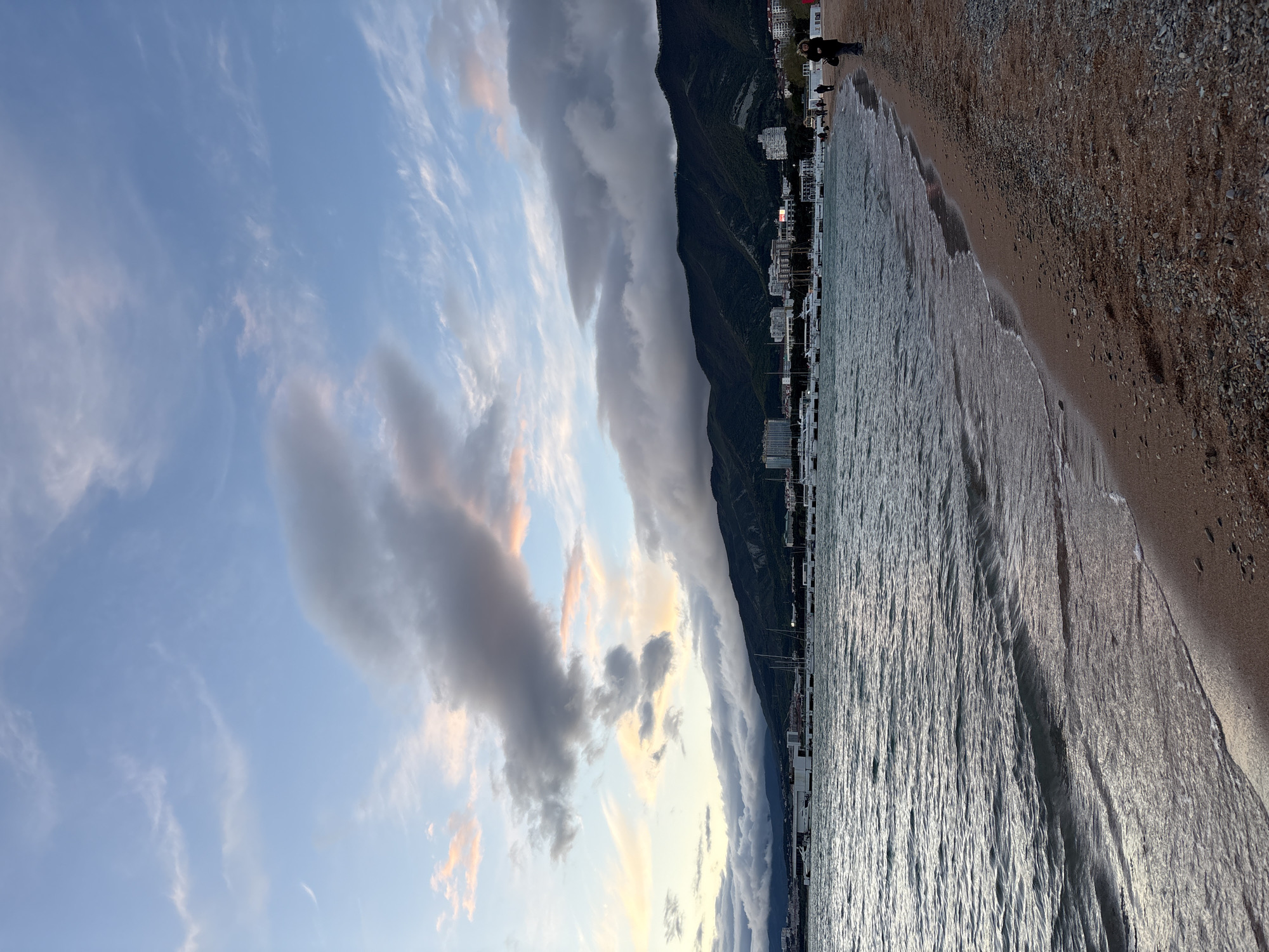 The image depicts a beach scene in Gelendzhik, Russia. The Black Sea is on the right side of the image, with waves gently lapping onto the sandy and pebbly shore. Adjacent to the beach are buildings and structures lining the coastline, interspersed with greenery, possibly trees or shrubs. A mountain range runs parallel to the coastline, further inland. A bank of clouds hangs over the mountains, some extending across the sky towards the left. Some people are visible walking along the beach. The sky features a mix of blue and lighter, scattered clouds, with hints of sunset hues.