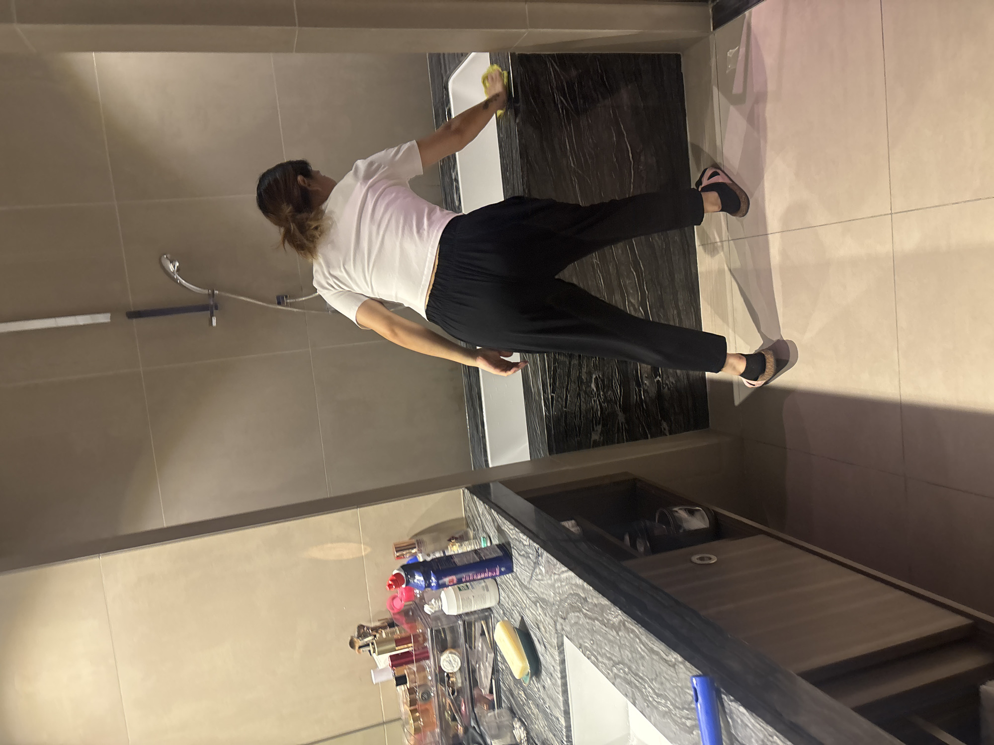 In a modern bathroom, a woman with dark hair pulled back in a ponytail is cleaning the edge of a bathtub. She is wearing a white crop top and black pants. The bathroom features large, light-colored tiles on the walls and floor, with a dark, marble-patterned surface around the bathtub. A sink and vanity with various toiletries are visible to the left. The scene takes place in Dubai, United Arab Emirates.