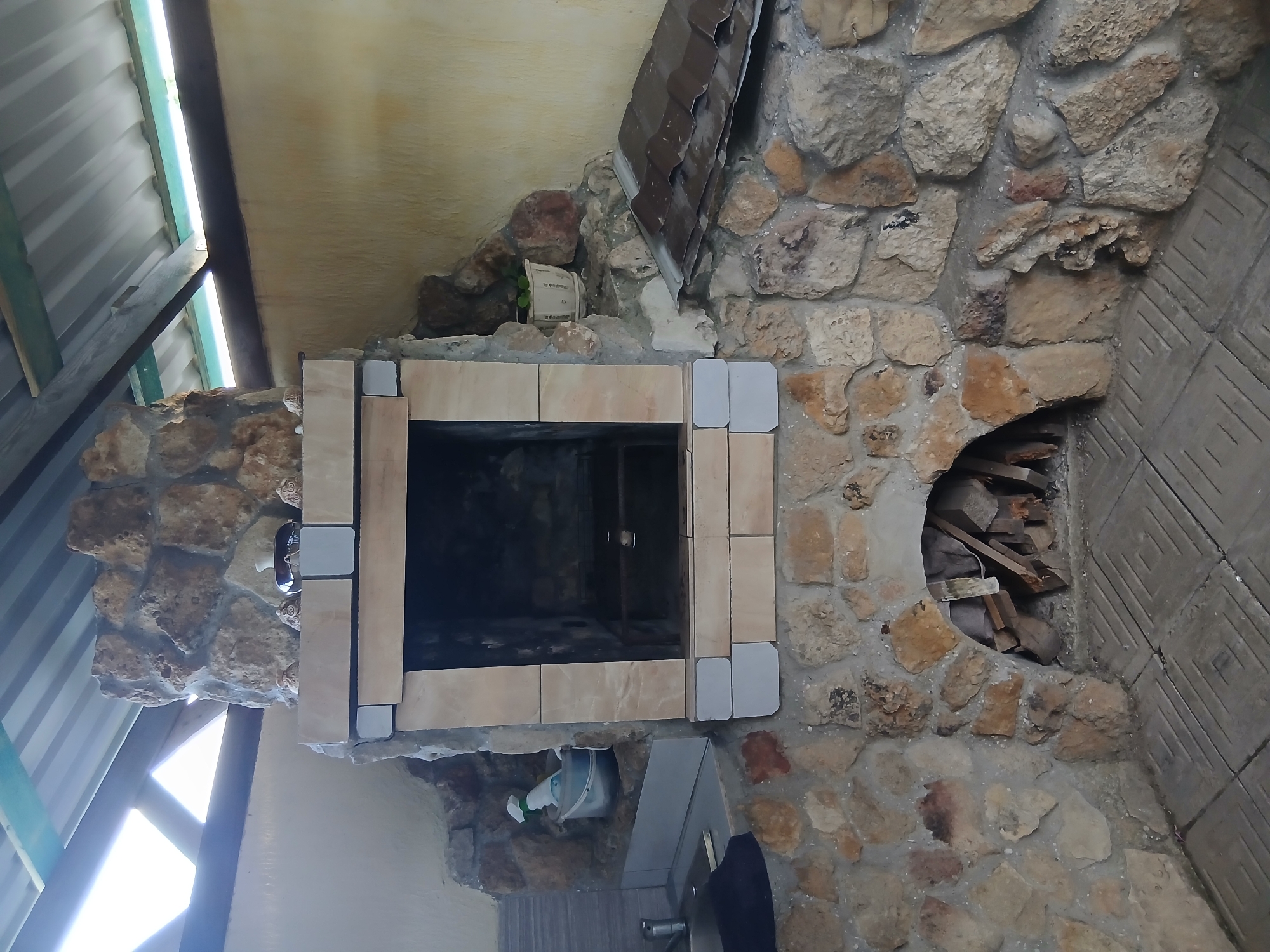 An outdoor stone barbecue with a tiled hearth is shown in this medium shot. The barbecue features a large rectangular opening with light-colored stone accents and a metal grate. Above the opening, a vase and several decorative objects are displayed on a stone ledge. To the left of the barbecue is a small sink area with a faucet and a bucket containing a spray bottle. To the right, a small potted plant sits on a stone ledge. The barbecue is situated under a corrugated metal roof supported by wooden beams. The ground is covered with square tiles. The overall scene suggests an outdoor kitchen or dining area. The location is identified as Unknown, Unknown.