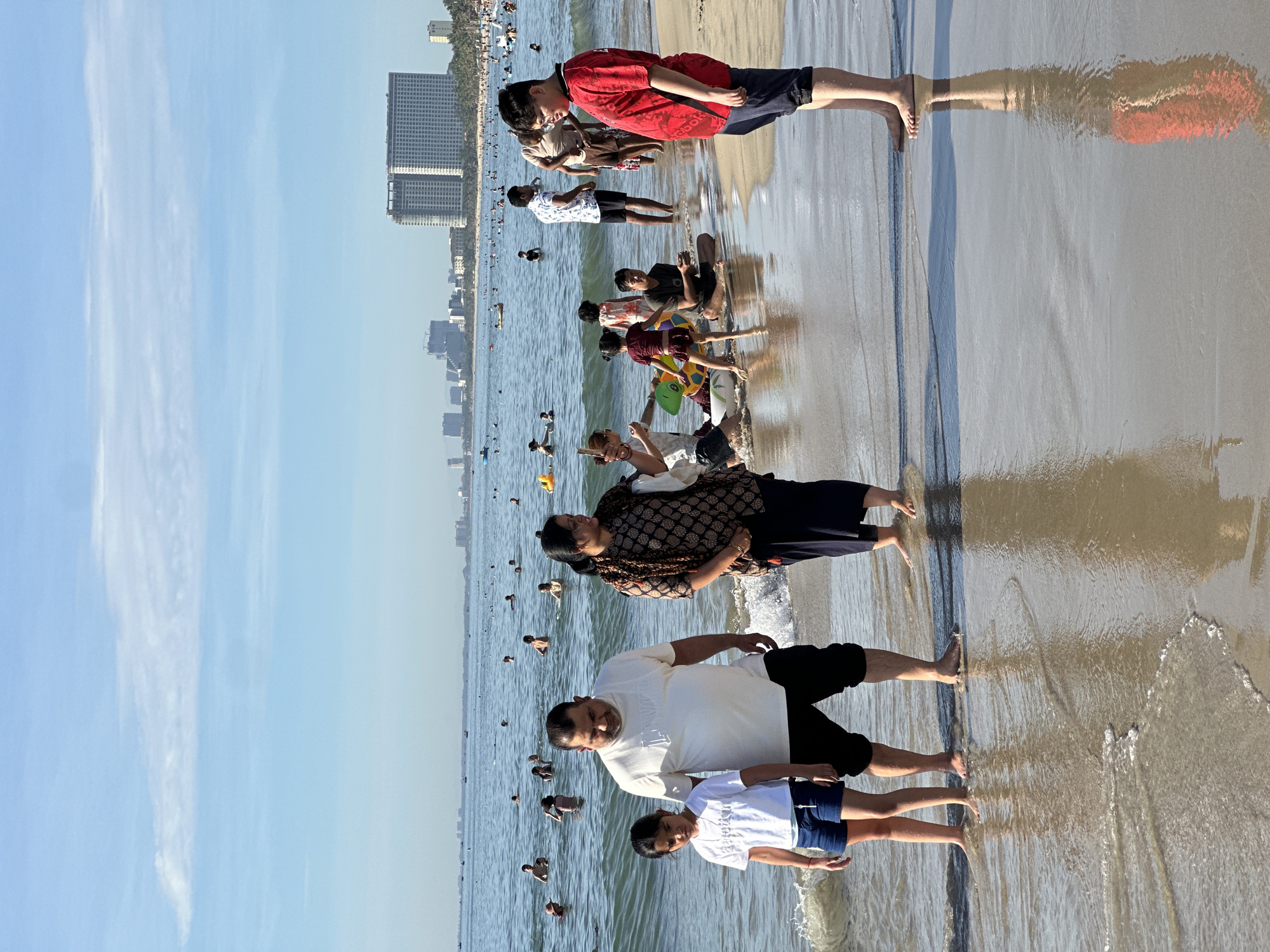 A sunny day at My Khe Beach in Da Nang, Vietnam, shows numerous people enjoying the ocean and shore. In the foreground, a family of three stands near the water's edge. A young girl in a white t-shirt and blue shorts stands to the left, looking down. Beside her, a man in a white t-shirt and black shorts gazes forward. To the right of the man, a woman in a patterned top and dark skirt looks off to the side. Further back, other beachgoers are visible in the shallow water and on the sand. Some people are swimming, while others are wading or sitting. Children are playing with inflatable toys, including a turtle-shaped float. In the background, the cityscape of Da Nang is visible with high-rise buildings lining the coast. The ocean is calm with gentle waves rolling onto the sandy beach, which reflects the sunlight and the figures on the shore. The sky is a clear blue with a few wispy clouds. Boats are dotted in the distance on the water.