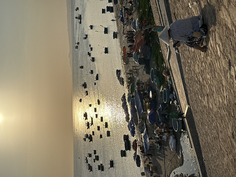 The image displays a coastal scene at sunrise or sunset in Thành phố Phan Thiết, Vietnam. The sun, a bright disk, is high in a hazy, golden-orange sky, casting a strong reflection across the calm ocean. Numerous fishing boats, including smaller circular basket boats, are scattered across the water, many appearing as silhouettes against the sun's glare.
On the sandy beach, a market operates with multiple temporary stalls and shelters, identifiable by various colored tarps and umbrellas (blue, green, orange, striped). Numerous individuals are gathered around these stalls, indicating commercial activity. Large circular basins are present on the sand near the market area.
In the foreground, a textured, sloped stone or brick embankment descends towards the beach. A person wearing a traditional conical Vietnamese hat (nón lá), a light-colored long-sleeved shirt, and patterned pants sits facing away from the viewer on this embankment. They are holding and observing a smartphone. A small woven bag is slung over their shoulder, and a clear plastic cup rests nearby. To the left, another person in a green shirt walks down the embankment. A third individual, possibly a child, stands near the seated person on the right.
