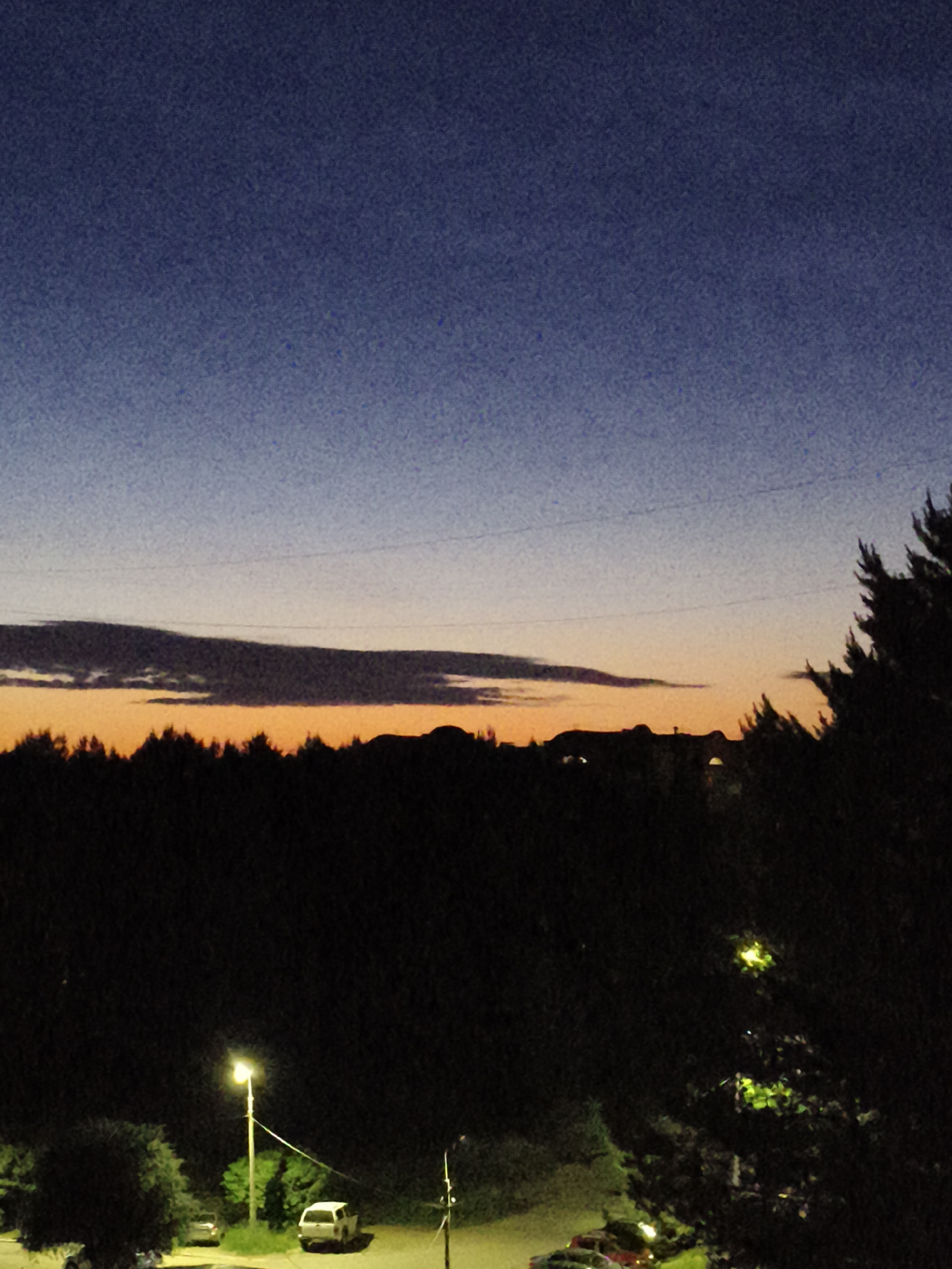 This image depicts a dusk or dawn scene in an unknown urban or suburban location. The sky occupies the upper two-thirds of the frame, presenting a gradient from dark indigo-blue at the top, transitioning through a lighter blue-grey to a prominent horizontal band of orange-yellow light along the horizon. A long, dark, horizontal cloud formation is silhouetted against this bright band, with thinner, dark power lines faintly visible above the clouds.
The horizon is characterized by a dense, dark silhouette of coniferous trees. Above this tree line, particularly towards the right, the indistinct silhouettes of distant buildings or structures are visible.
The lower third of the image is largely dark, illuminated by artificial light sources. A bright, yellow-glowing streetlight mounted on a white pole stands in the lower-left quadrant, illuminating a paved area and surrounding green foliage. A white SUV is clearly parked beneath this streetlight, with another darker vehicle partially visible to its left. Towards the lower-right, the tops of additional parked cars, including a red one, are discernible amidst dark trees and shrubs. The entire image exhibits a noticeable grain and noise texture, indicative of low-light photographic conditions. No individuals are visible.
