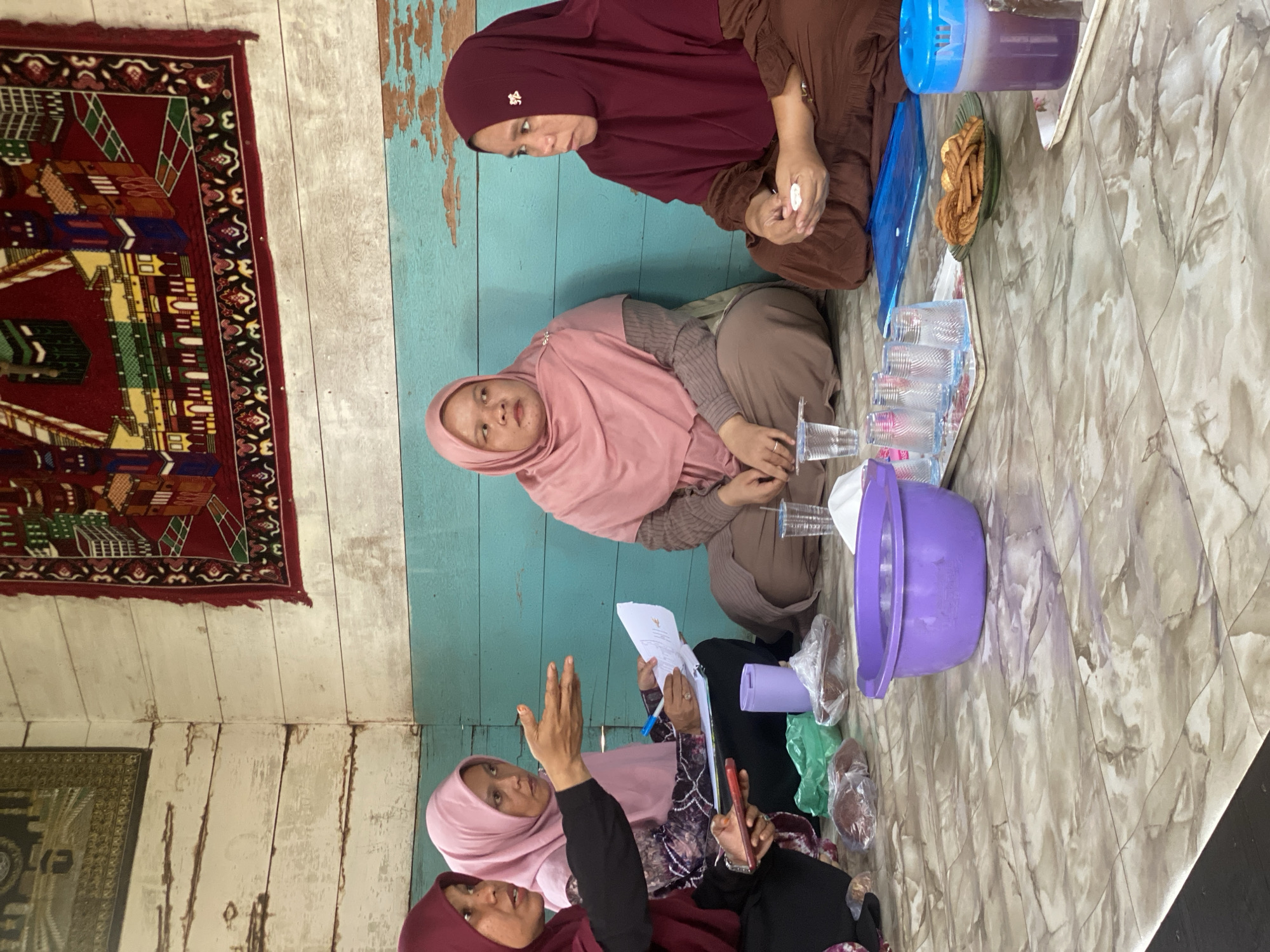 Four women, dressed in hijabs and modest clothing, are seated on a patterned floor in what appears to be a room with wooden walls. The walls are painted with horizontal planks, with a section of turquoise-colored wood above white-washed planks. A large, decorative tapestry featuring a depiction of buildings and intricate patterns hangs on the upper portion of the wall.
The women are gathered in a semi-circle. The woman on the far left, wearing a maroon hijab and black top, is actively gesturing with her right hand and holding a red mobile phone in her left. To her right, a woman in a pink hijab is looking towards the gesturing woman while holding a piece of paper and a pen. The third woman, also in a pink hijab, is seated further back and looking slightly to her left. The fourth woman, on the far right, wears a maroon hijab and a brown dress, and is looking downwards at her hands.
On the floor in front of the women are various items, including a purple bowl filled with water, plastic bags containing food items, plastic cups, a small plate of crackers, and a jug with a blue lid containing a purple liquid. A blue tray is also visible. The overall impression is that of an informal gathering or meeting. The specific location in Bawahan Selan, Indonesia, is not visually discernible from the image.