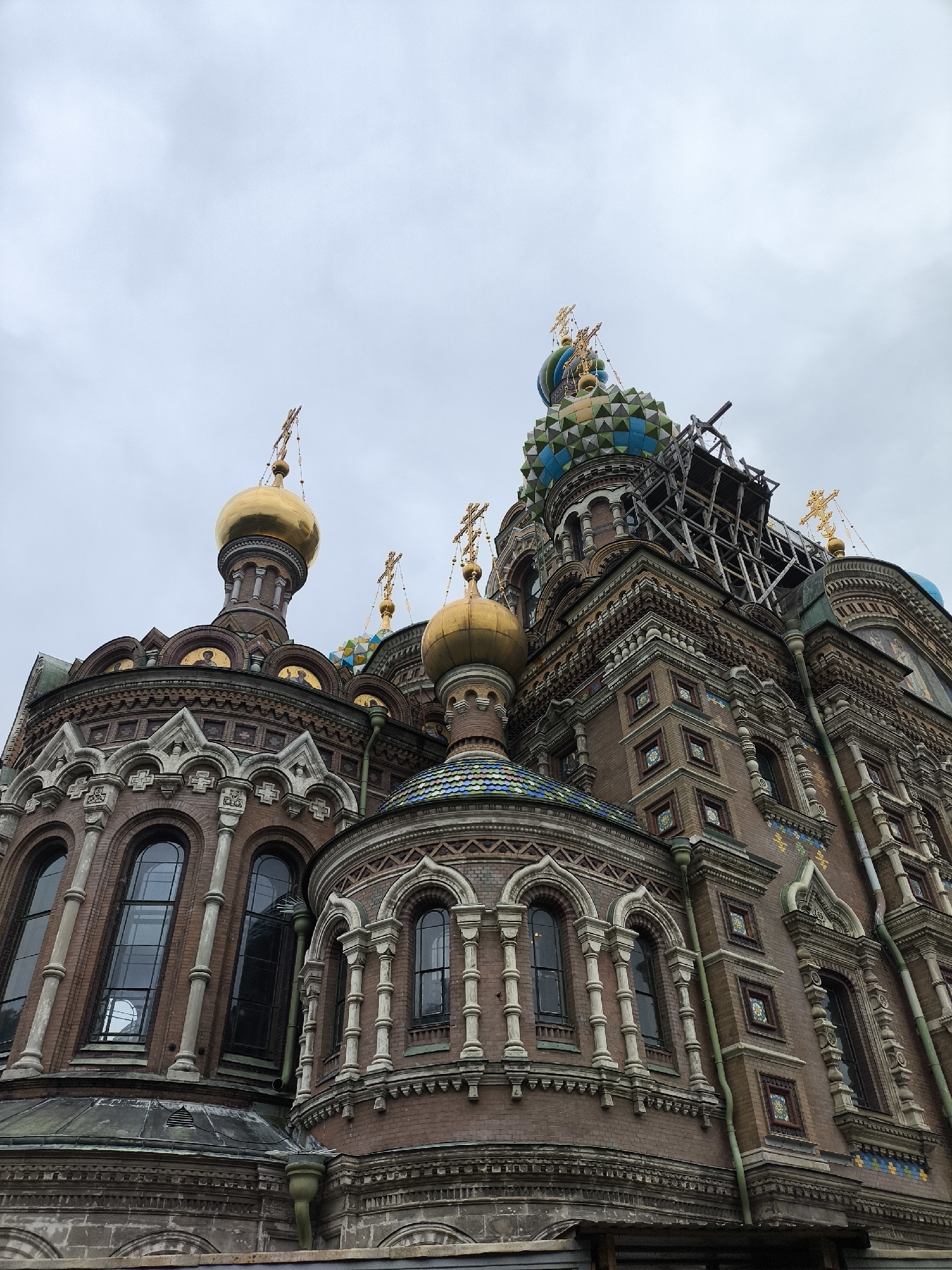This media file shows a low-angle, exterior view of the Church of the Savior on Spilled Blood in Saint Petersburg, Russia. The church features multiple onion domes, some of which are gold-plated and topped with crosses, while others are adorned with intricate mosaic tiles in shades of blue, green, and yellow. The facade is constructed of red brick with ornate decorative elements, including arched windows, columns, and patterned tiles. Scaffolding is visible around one of the domes, suggesting ongoing maintenance or restoration work. The sky is overcast, indicating a cloudy day. The image captures the architectural grandeur of the church, highlighting its distinctive Russian Revival style. No people are visible in the image.