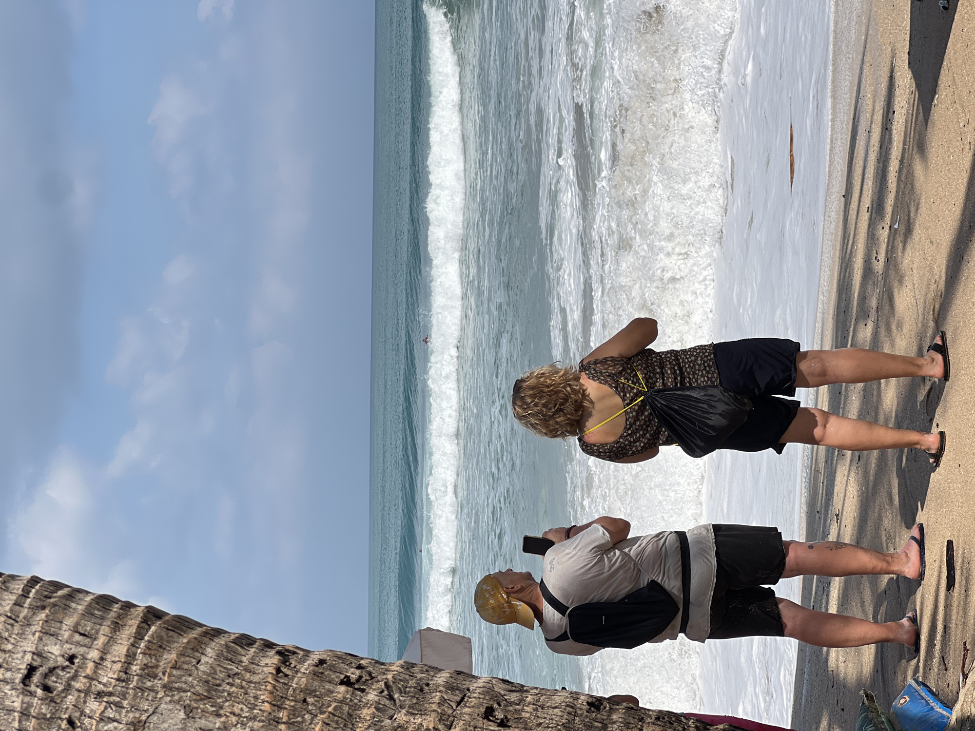 Two individuals, a man wearing a yellow cap and a woman with curly hair, stand on a sandy beach, facing a choppy ocean with large waves. The man, on the left, is holding a smartphone and appears to be taking a picture or video. He is wearing a light-colored t-shirt, black shorts, and a black backpack. The woman, on the right, is wearing a patterned top and dark shorts, also with a black backpack. In the distance, a small figure, possibly a surfer, can be seen in the ocean. A portion of a palm tree trunk frames the left side of the image. The scene appears to be located in Kuta, Indonesia, a popular tourist destination known for its beaches and surf.
