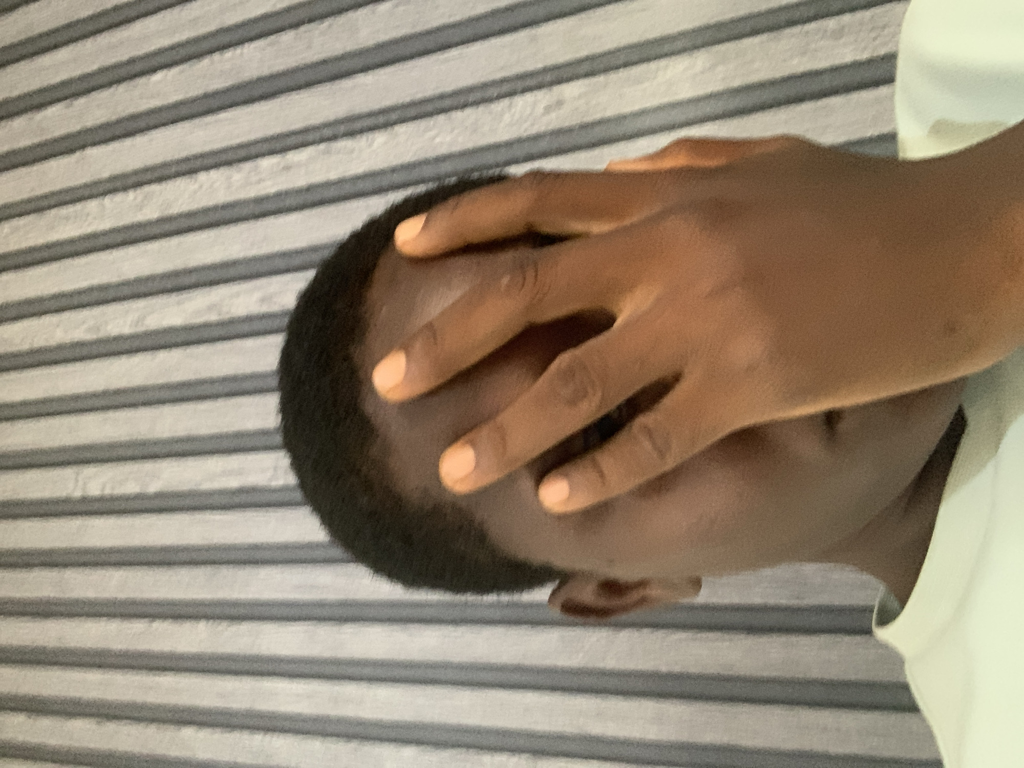 A close-up, high-angle shot shows a person's face and hand in the foreground, with a textured wall in the background. The person has dark skin and short dark hair. Their right hand is covering their face, with the fingers spread. The person is wearing a light-colored t-shirt. The background wall has vertical stripes of alternating light gray and white. The image is taken in Jos, Nigeria.