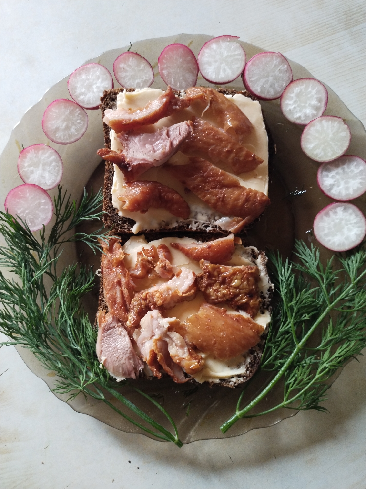 A top-down photographic depiction of a meal arranged on a translucent dark plate, located in Horishni Plavni, Ukraine. The plate contains two open-faced sandwiches, each featuring a slice of dark bread spread with a pale yellow substance (likely butter or cream cheese), topped with irregularly shaped pieces of cooked meat, ranging in color from light pink to golden-brown and reddish-brown, indicative of poultry or smoked fish. Fresh green dill sprigs are arranged in an arc along the bottom and left side of the plate, while thin, round slices of radish, displaying white flesh with pink-red edges, are positioned symmetrically along the top and right side. The plate, which has an irregular, wavy rim and shows faint moisture residue, rests on a light-colored surface, possibly a wooden table. No people are visible in the scene.
