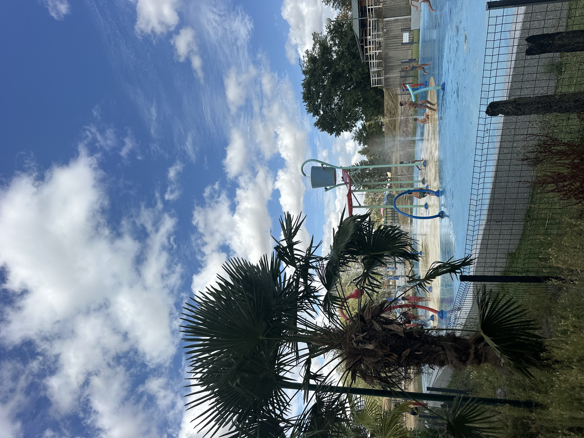 A bright, sunny day with scattered clouds over Jablines, France. In the foreground, a palm tree with large fronds stands to the left, obscuring part of the scene. A black chain-link fence runs horizontally across the lower third of the image, separating the viewer from the main subject. Beyond the fence is a shallow splash pad with bright blue flooring. Several children are playing in the water. Some are running, while others are standing near water features that spray jets of water. A large blue bucket, a staple of splash pad attractions, is visible on a metal structure in the background. To the far right, a wooden building with a white railing overlooks the splash pad. Trees and foliage are visible in the background, suggesting a park or recreational area.