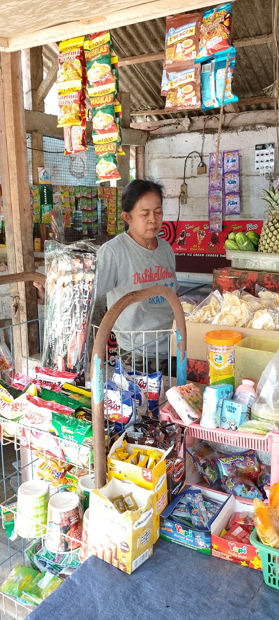 The image depicts a small, open-air convenience store in Pekutatan, Indonesia. A middle-aged woman, with her eyes closed and head slightly bowed, stands behind a counter. She is wearing a grey t-shirt with the text "Dislike Bohr" and "OKANA" visible. The store is packed with a variety of packaged goods, including snacks, drinks, and household items. Packaged snacks hang from the ceiling, such as yellow and red packets with Indonesian text and images of food. On shelves behind the woman are rows of colorful packaged goods. In front of her, on the counter and in wire racks, are more packaged snacks, drinks, and various food items like chips and crackers. A pineapple and some cucumbers are visible on a shelf to the right. The overall impression is a modest, well-stocked local shop.