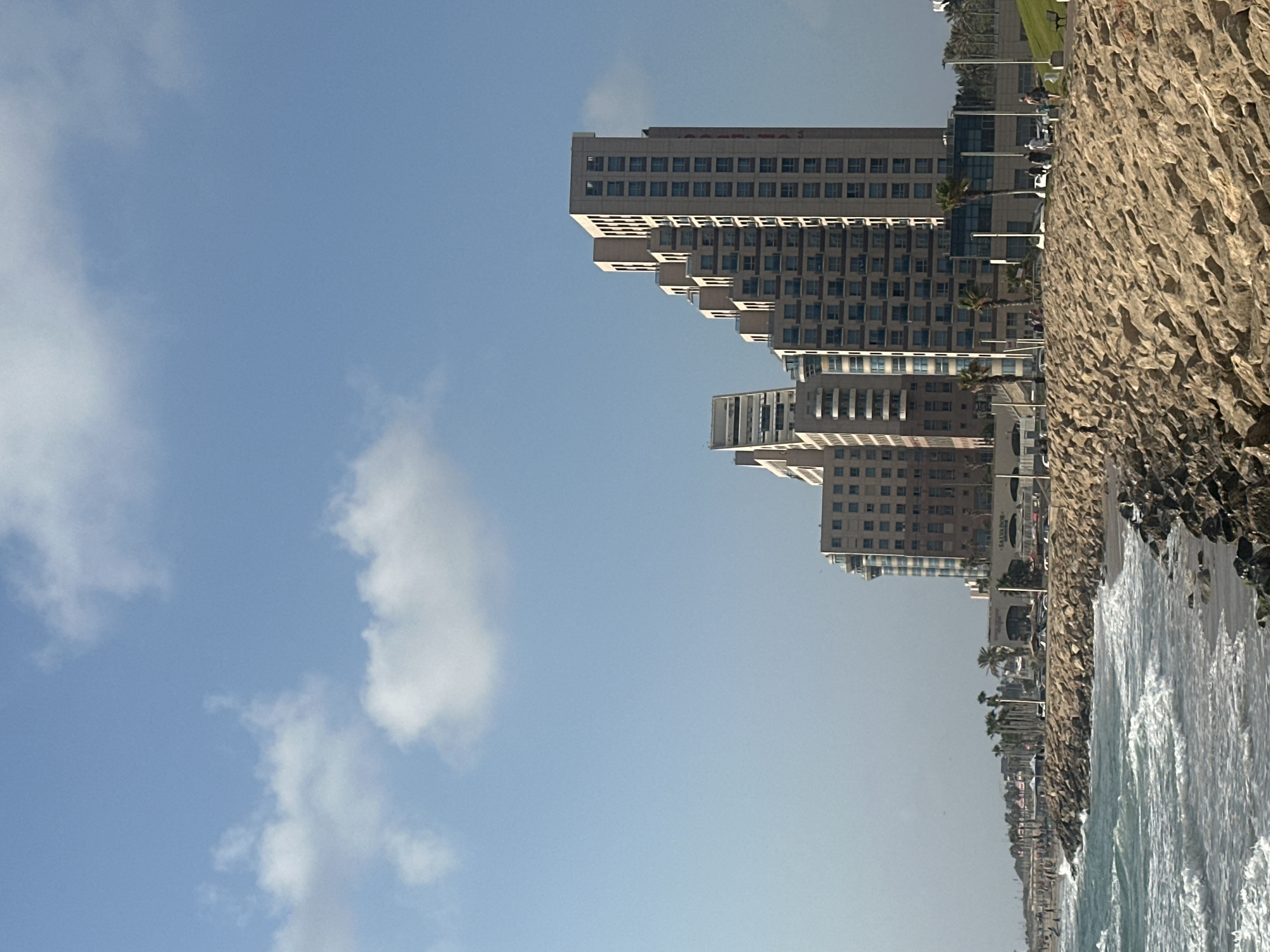 The image shows a seaside view in Haifa, Israel, with buildings and the Mediterranean Sea. A rocky breakwater lines the shore, with waves crashing against it. Behind the breakwater, a sandy beach is visible, with people walking and sitting. Several tall buildings, some with a stepped design, stand on the coast. Palm trees are scattered along the coastline, and the sky is blue with a few white clouds. The overall scene depicts a sunny day at a coastal urban area.