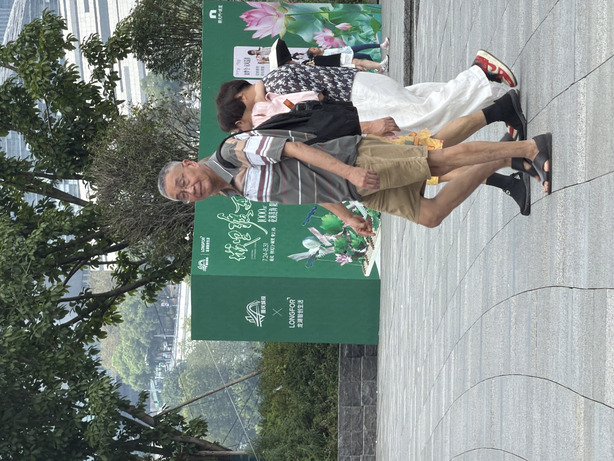 This is a medium shot image taken outdoors in Yuzhong District, China, of several people walking. The foreground shows a paved walkway made of large, gray rectangular tiles with curved dark lines between them. In the mid-ground, an older East Asian man with glasses and gray hair is walking towards the right, wearing a striped polo shirt and khaki shorts. He has a backpack on his left shoulder. To his right, another person, partially obscured, is walking with their arm around the man's shoulders, wearing a white outfit and a patterned shirt over a pink garment. Behind them, a green billboard is visible with Chinese text and images of lotus flowers and people. In the background, trees and modern buildings can be seen. The overall lighting suggests it is daytime.