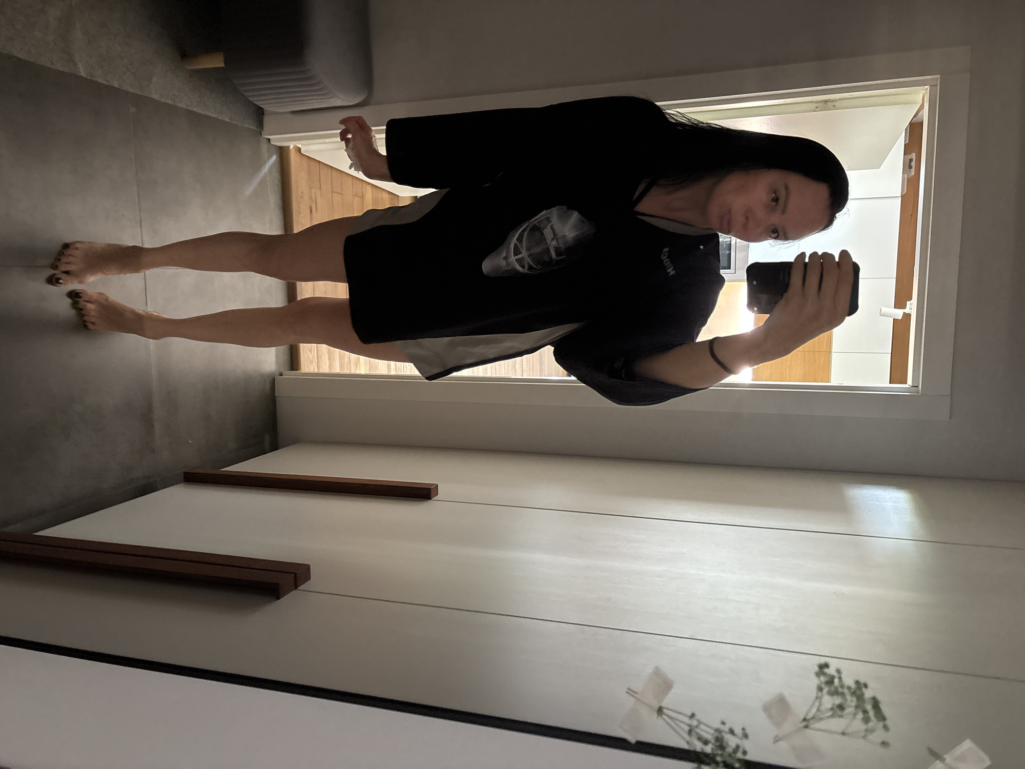 A woman is taking a selfie in front of a mirror in an indoor setting. She has dark hair and is wearing a black shirt with a print on the front, and short gray shorts. She is holding a smartphone in her right hand, and the camera is facing her. Her feet are bare. The mirror reflects the rest of the room, including a cabinet with brown handles, a plant in a vase, and an open doorway to another room with a gray sofa. The setting appears to be a modern apartment. The location is likely Chaikovsky, Russia, based on the image source data.