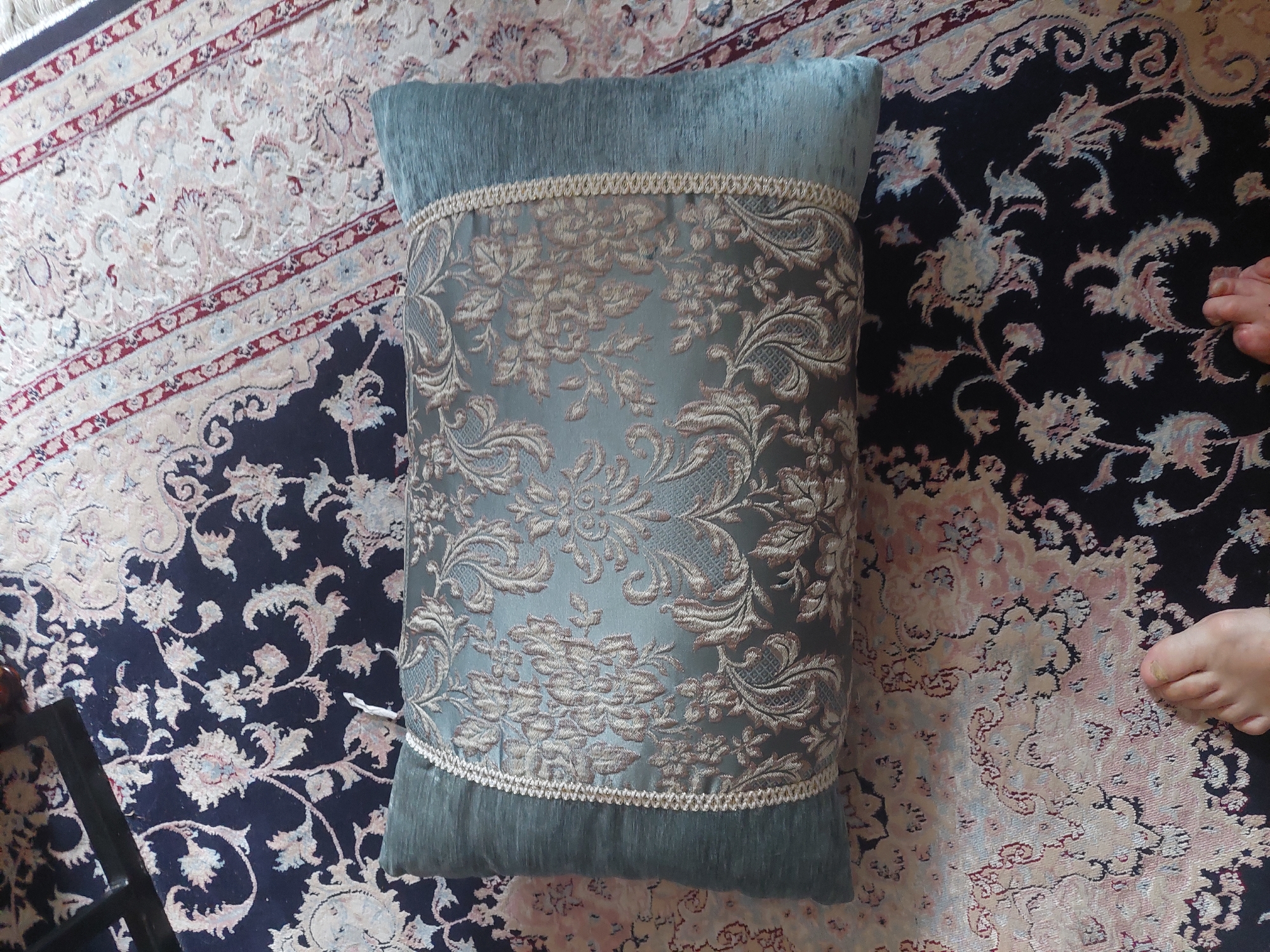 A rectangular, teal pillow with ornate gold embroidery and a textured border lies on a dark blue Persian rug with a floral pattern. A portion of a human foot, with visible toes and a toenail, is visible in the lower left corner of the image, resting on the rug. The scene is indoors, likely in a home. The location is unknown and cannot be determined from the provided image.