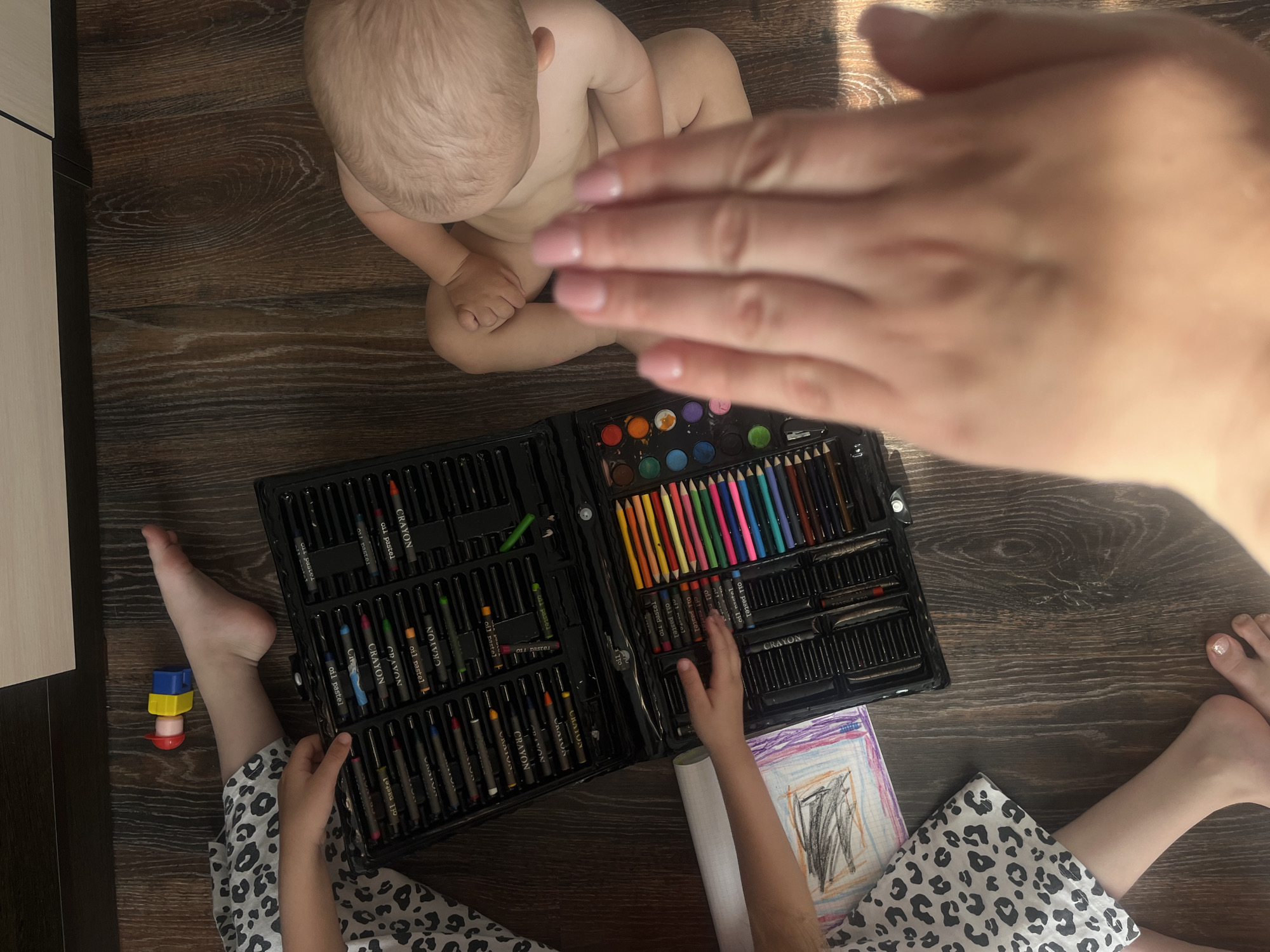 In an indoor scene on a wooden floor, a young child, partially visible from the torso up, sits with their legs crossed. To the left, another child's legs and feet are visible, one foot resting near a set of colorful crayons and drawing supplies in a black case. A child's drawing is also present. A toy with red, yellow, and blue components lies on the floor near the child's foot. The scene is illuminated by natural light, casting shadows on the floor.