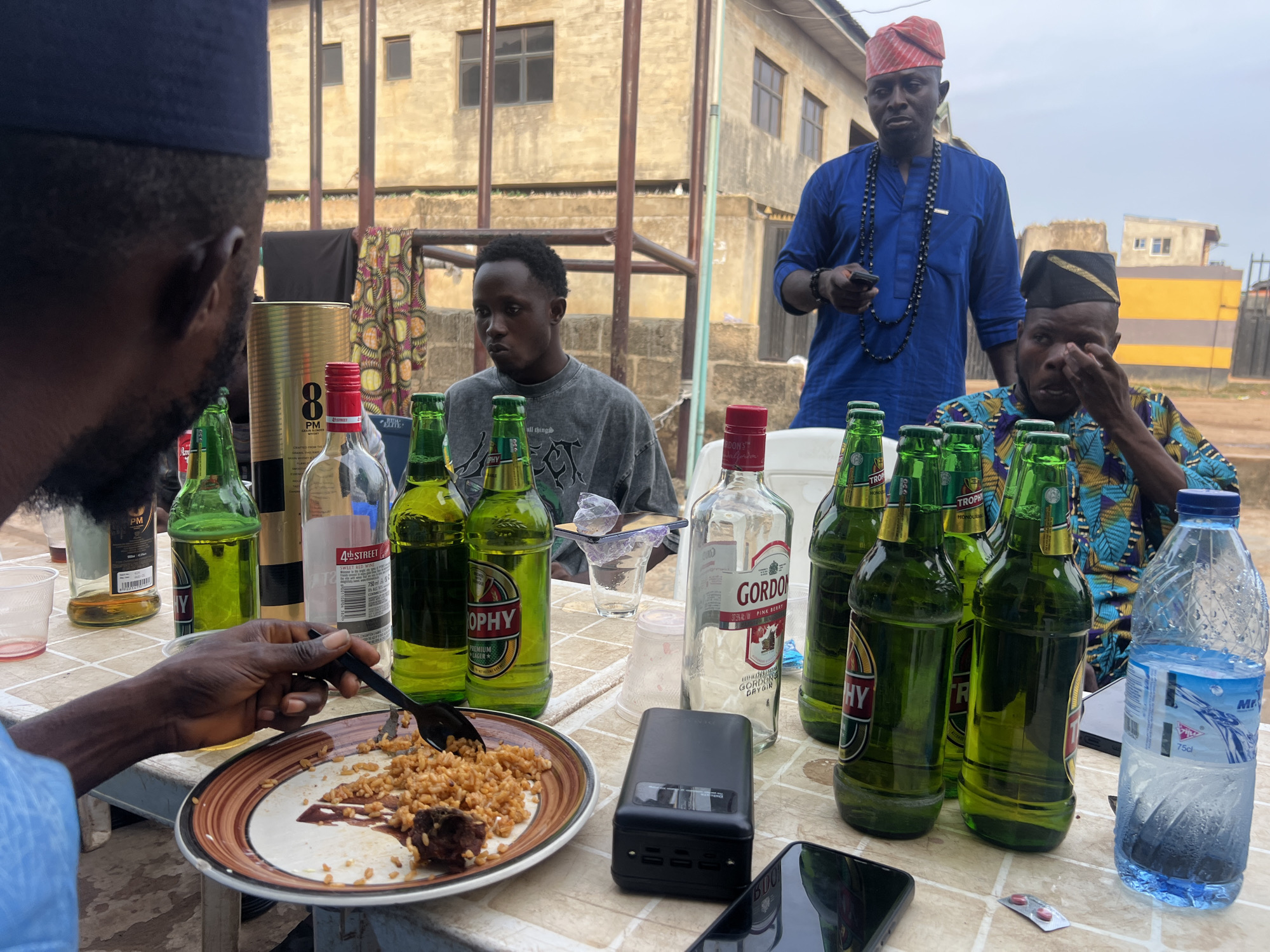 In Ota, Nigeria, a group of men are gathered around a table laden with food and drinks. The scene appears to be outdoors, possibly on a balcony or patio, with a building in the background. The men are dressed in a mix of traditional and casual attire. One man, wearing a blue shirt and a red cap, stands and holds a mobile phone. Another man, seated, is wearing a patterned shirt and appears to be touching his eye. A third man, in a grey t-shirt, is seated and looking down. On the table, there are several bottles of beer, a bottle of Gordon's Pink Gin, and a bottle of water. A plate of rice with meat is being eaten by one of the men, who is using a black spoon. There are also plastic cups and a power bank on the table. The overall atmosphere seems relaxed and social.