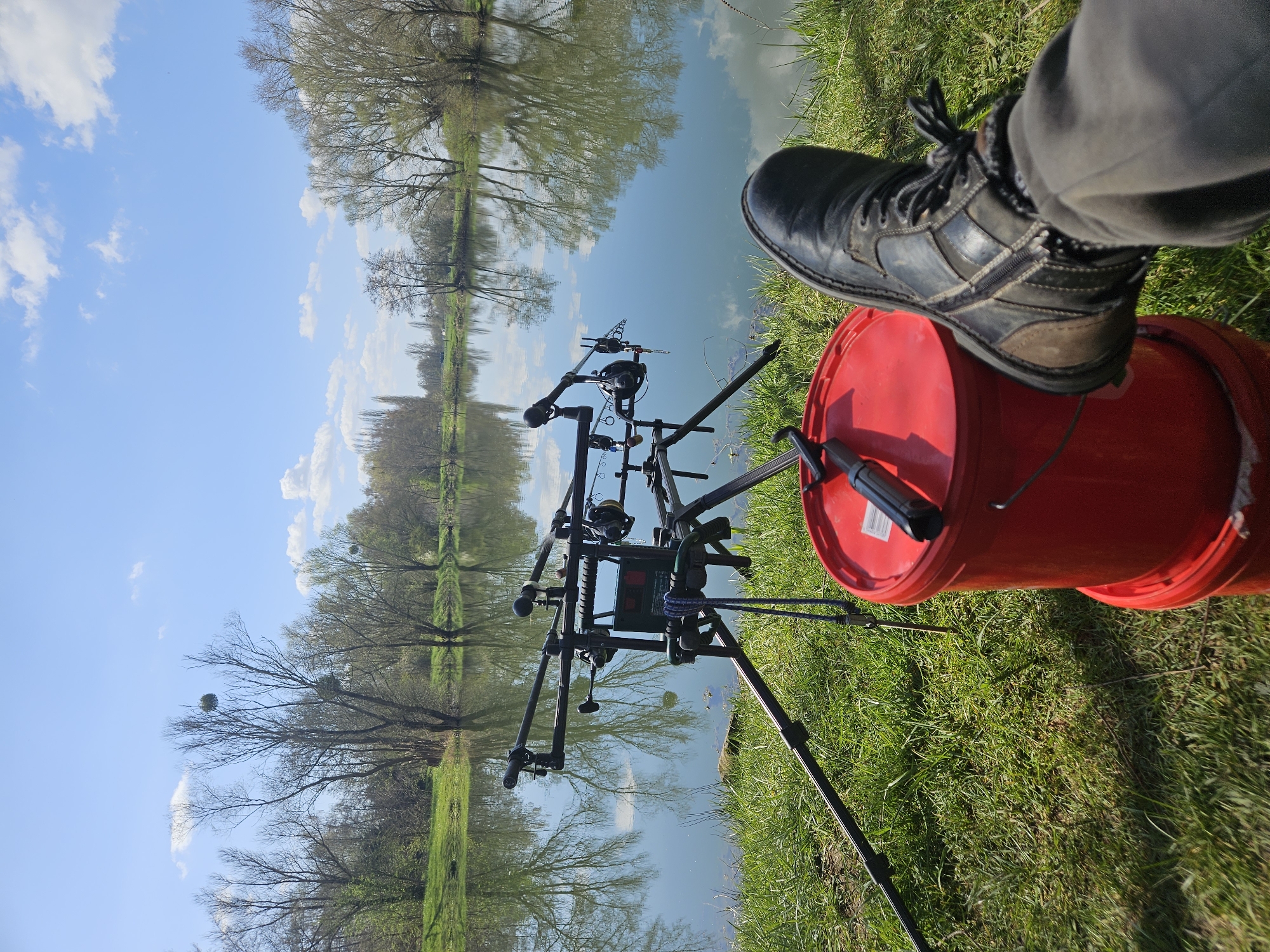 A fishing scene unfolds in Gavisse, France, beside a tranquil body of water reflecting the sky and surrounding trees. A fishing rod stand, equipped with multiple fishing rods, sits poised at the water's edge. Adjacent to the stand, a red bucket is positioned on the grass. A person's leg and boot rest atop the bucket.
