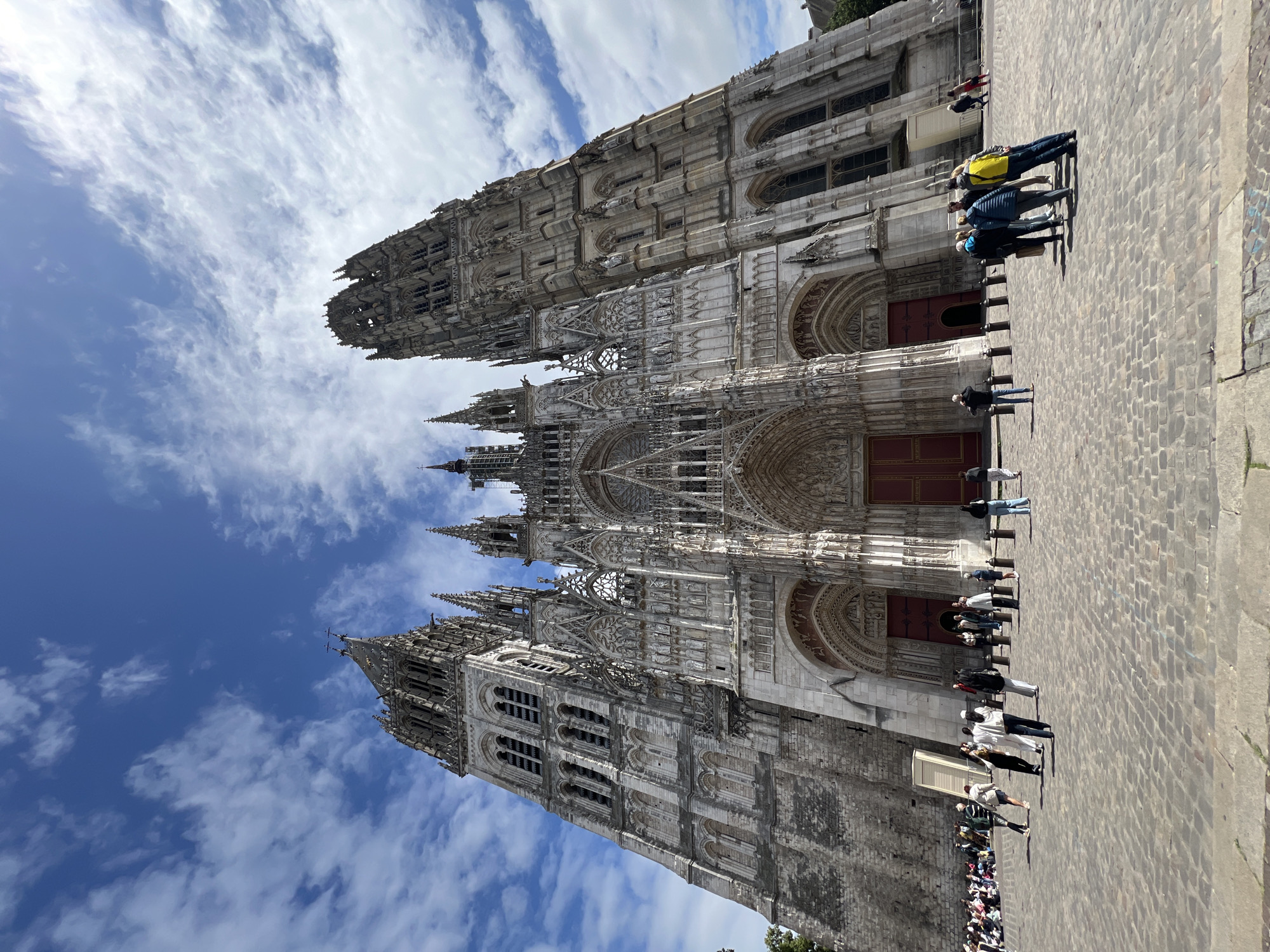 The image set displays the magnificent Gothic facade of Rouen Cathedral in Rouen, France. The grand cathedral dominates the scene, showcasing its elaborate architecture, including multiple towering spires, intricate stone carvings, pointed arches, and decorative rose windows. Several ornate doorways, some featuring prominent red doors, are visible at the ground level entrances.
In front of the cathedral stretches a wide, paved cobblestone square. Numerous individuals and small groups of people are scattered across this plaza. Many appear to be tourists or visitors, standing, observing the monumental structure, or congregating. Their distinct, elongated shadows on the ground indicate bright sunlight, suggesting a clear daytime setting. The sky above is a mix of vibrant blue and scattered white, wispy clouds, contributing to the fair weather impression.
The detailed craftsmanship of the cathedral, from its complex sculptural elements to its layered buttresses and pinnacles, is a key visual feature. No visible text or signage is discernible on the building itself or within the plaza. The scene captures the awe-inspiring presence of the historic landmark amidst visitor activity.