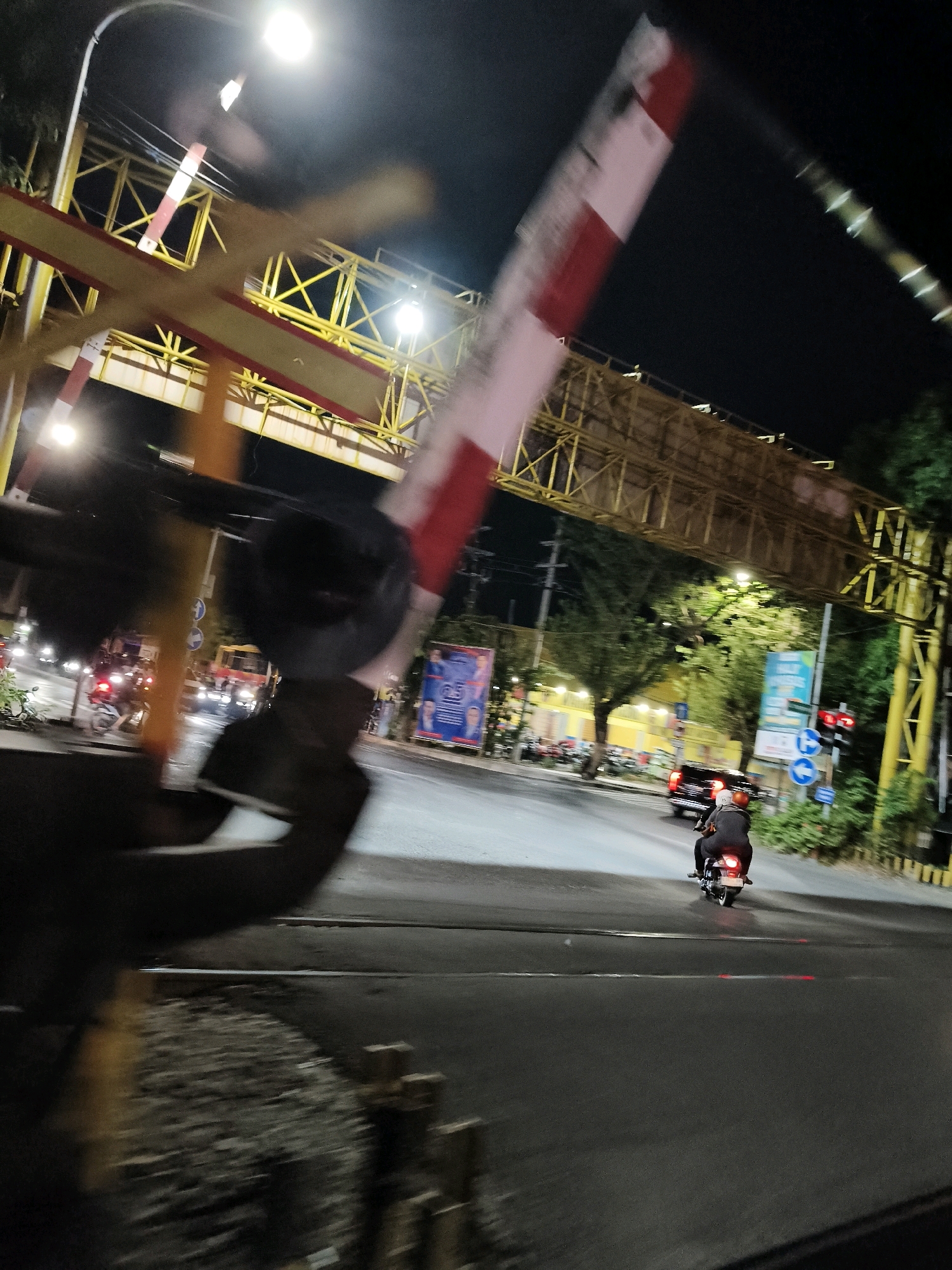 The image captures a night scene at a railway crossing in Surabaya, Indonesia. A railway crossing barrier, painted red and white, is prominently displayed in the foreground, blurred due to motion. Beyond the barrier, the street is illuminated by streetlights and vehicle headlights. A yellow metal structure, possibly an overpass or a sign frame, spans across the upper part of the image. In the middle ground, traffic is visible, including a black car and several motorcycles. One motorcycle, carrying two individuals, is moving away from the camera. On the right side of the street, there are trees and a traffic light. Billboards are also visible in the background, one of which displays an image of two men and text in Indonesian. The overall atmosphere suggests a busy urban environment at night, with the railway crossing as a focal point.