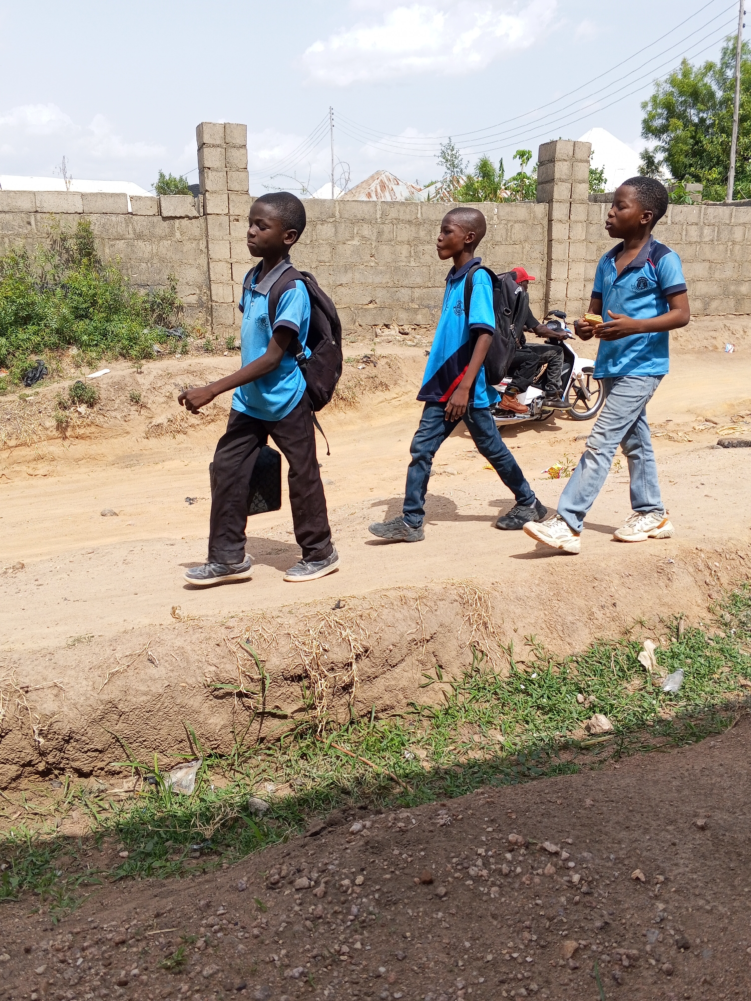 The image depicts three young boys walking along a dusty, unpaved road in Adonkolo, Nigeria, under a bright, partly cloudy sky. All three boys wear light blue polo shirts, resembling school uniforms, each with an emblem or crest visible on the left chest. The boy on the far left wears black trousers and a black backpack, carrying a dark object in his left hand. The middle boy wears dark blue jeans and a black backpack. The boy on the right wears light-colored jeans and holds what appears to be crackers or biscuits in his hands.
The boys are walking in a line from left to right across the frame, along a raised dirt path bordered by a lower, grassy ditch. In the background, a concrete block wall extends horizontally, behind which trees, sparse buildings, and power lines are visible under the bright daylight. A person on a red motorcycle is partially seen behind the middle boy. The road is dry and sandy, indicating dry weather conditions. The boys' expressions are neutral, focused on walking, suggesting an ordinary daily activity, possibly commuting to or from school.