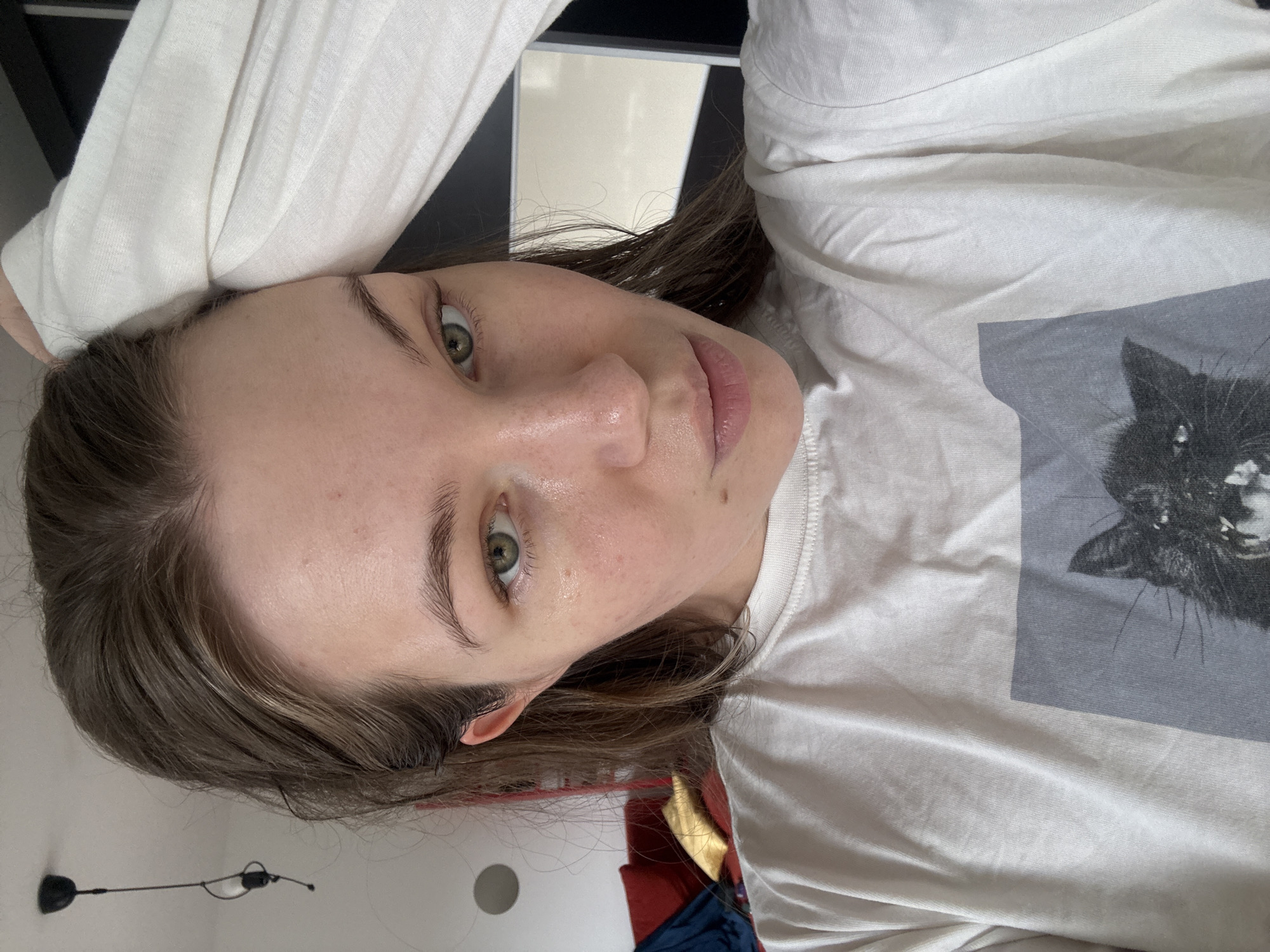 A woman with light skin, green eyes, and brown hair lies on a surface wearing a white long-sleeved shirt with a gray square design featuring a black cat's face. The location is indeterminate; no details suggest Vozdovac Urban Municipality, Serbia. The environment includes a wall with an indistinct object and a glimpse of other items.
