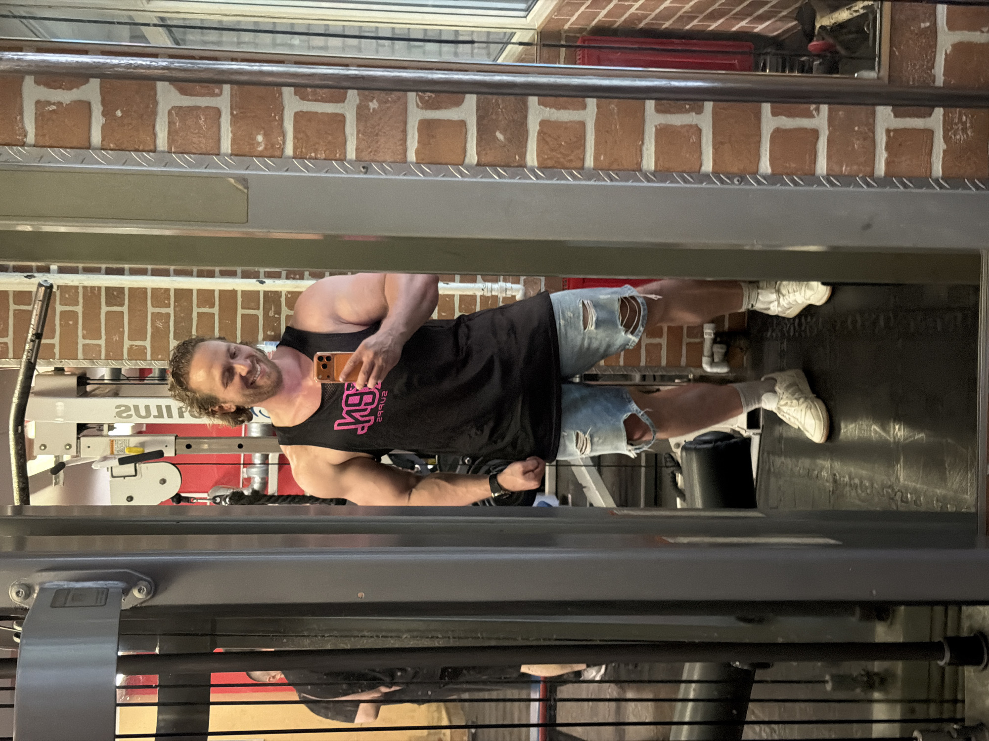 A muscular man with curly brown hair, wearing a black tank top with pink lettering and ripped denim shorts, stands in a gym, taking a selfie in a mirror. He is smiling and holding an orange smartphone. The gym has exposed brick walls and visible exercise equipment, including a weight machine and a large black weight bag on the floor. In the background, another person is visible in the reflection, wearing a black t-shirt. The image does not contain any information that would indicate the location as Saint Petersburg, Russia.