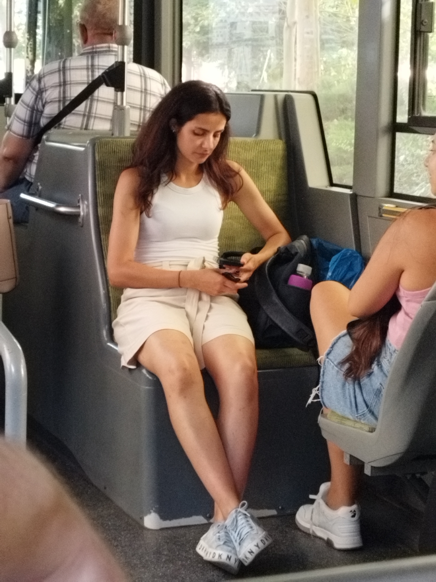 The image depicts an indoor scene, likely within a public transportation vehicle, possibly a bus or a tram. The lighting suggests daytime. The foreground features the legs and feet of two individuals. One person on the left is wearing light-colored sneakers with visible text that appears to be "DKNY". Their legs are crossed, and they are wearing a light-colored skirt. To the right, another individual is seated and wearing denim shorts and white sneakers. Their legs are extended.
In the mid-ground, a woman with long, dark hair is seated, wearing a white tank top and a light-colored skirt. She is holding a smartphone in her right hand and appears to be looking at it. Next to her is a black backpack containing a purple water bottle and a blue plastic bag. Behind her, an elderly man wearing a plaid shirt is seated, facing away from the camera.
The environment includes rows of seats, which are green and gray. There are window-like structures on the right side of the frame, revealing a blurred outdoor scene with greenery, suggesting trees. The floor is a dark, speckled material.
The specific location within the city of Bucharest, Romania, cannot be determined from the visual information provided in the image.