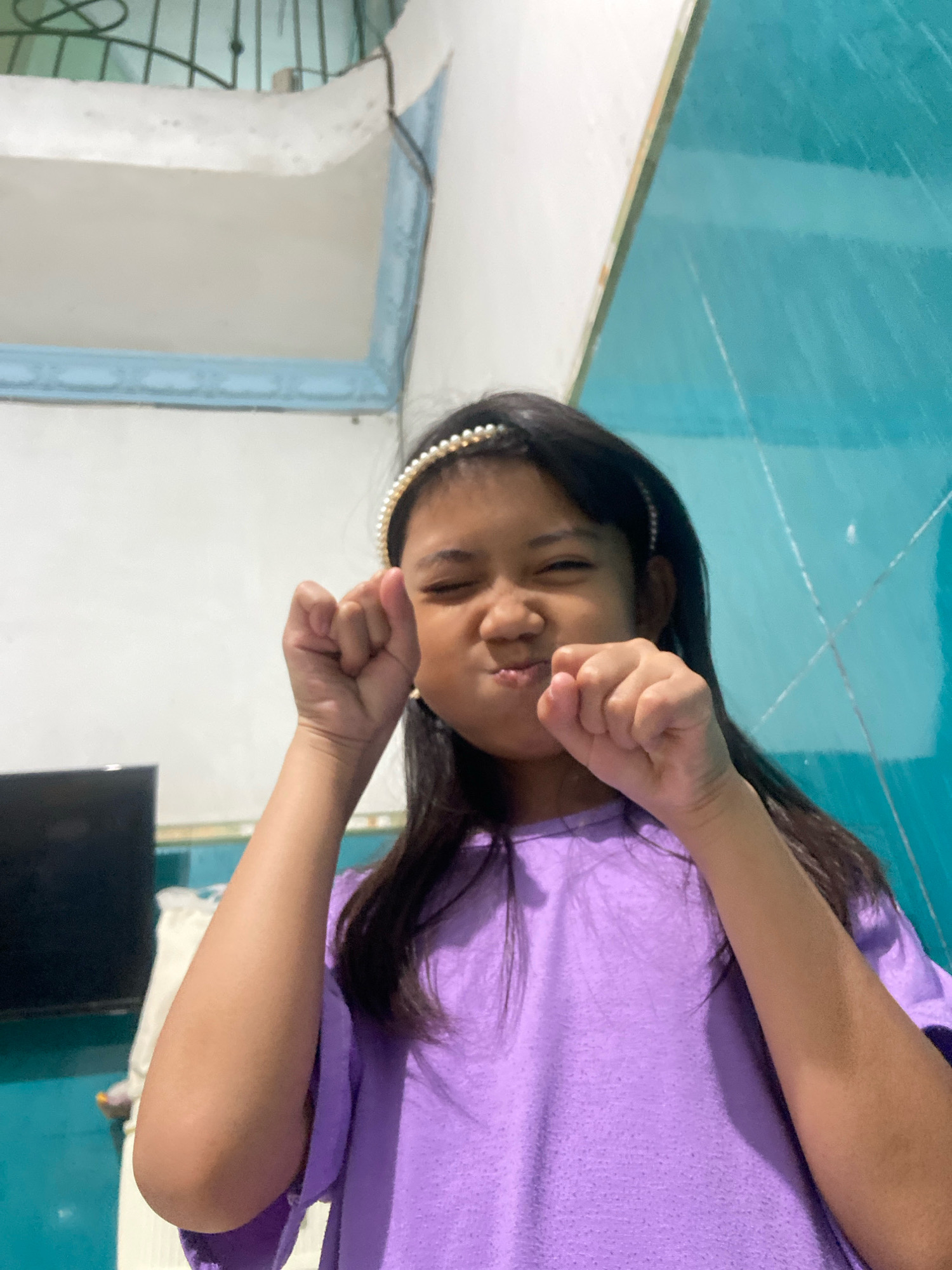 A young girl with dark hair and a pearl headband is posing for a photo. She is wearing a purple t-shirt and has her fists clenched near her face. Her eyes are closed and she has a playful expression. The background appears to be an indoor setting with a white wall on the left and a teal-colored tiled wall on the right. There is a window with bars visible in the upper left corner. A black television is visible on the far left. The image was taken in Gelam, Indonesia.