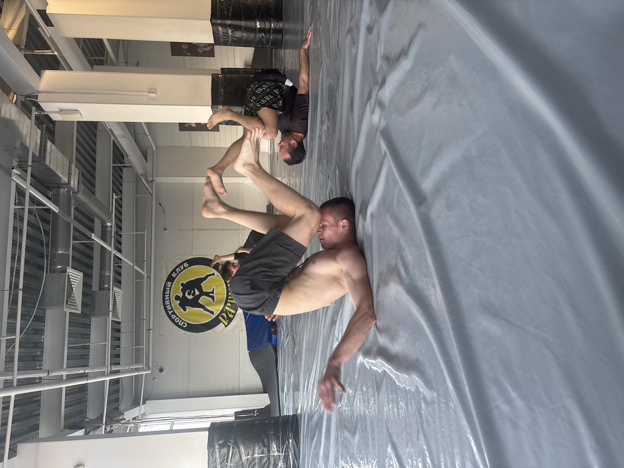 Two men are engaged in acrobatic wrestling on a gray wrestling mat in a gymnasium. One man is lying supine on the mat, supporting the other man on his feet, who is balancing upside down with his hands touching the mat. The location is likely a wrestling club in Perm, Russia, based on the Cyrillic script on a circular banner in the background that reads "СПОРТИВНЫЙ КЛУБ" ("Sport Club"). Gym lighting, ceiling fixtures, and supportive beams are visible in the background. There's also a glimpse of a person in a blue shirt and grey pants standing in the background. Black padded equipment is visible along the wall. The mat surface appears slightly wrinkled and reflective.